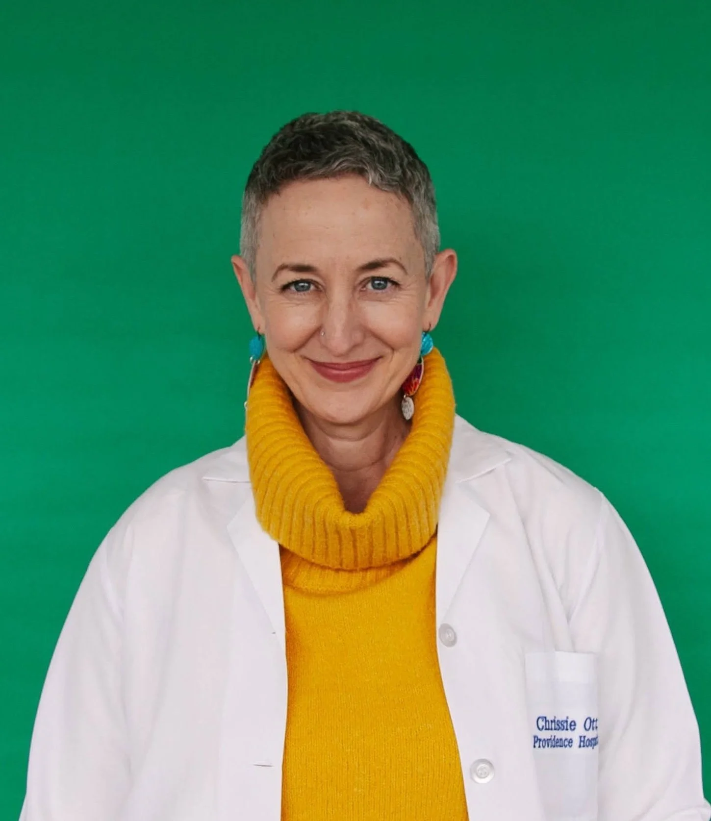 A woman with short gray hair, blue eyes, and a nose piercing, wearing a white coat over a yellow sweater with a turtleneck, standing against a green background.