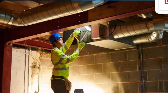 Construction worker in safety gear inspecting ductwork in a building basement.