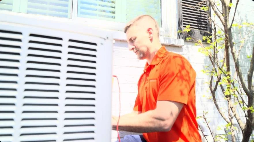 Person in orange shirt servicing outdoor air conditioning unit near a house.