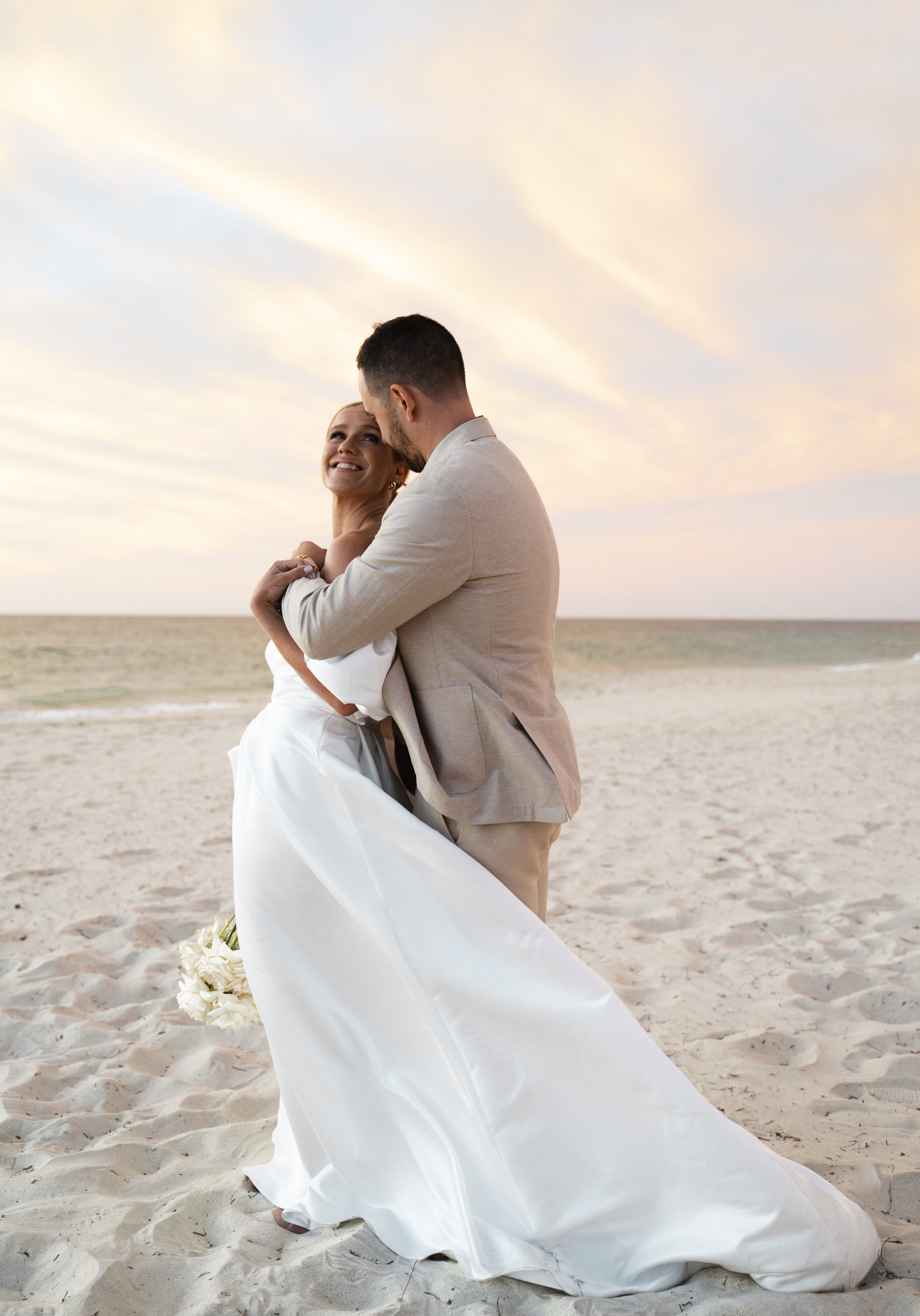 Couple in wedding attire embracing on a beach, sunset sky.