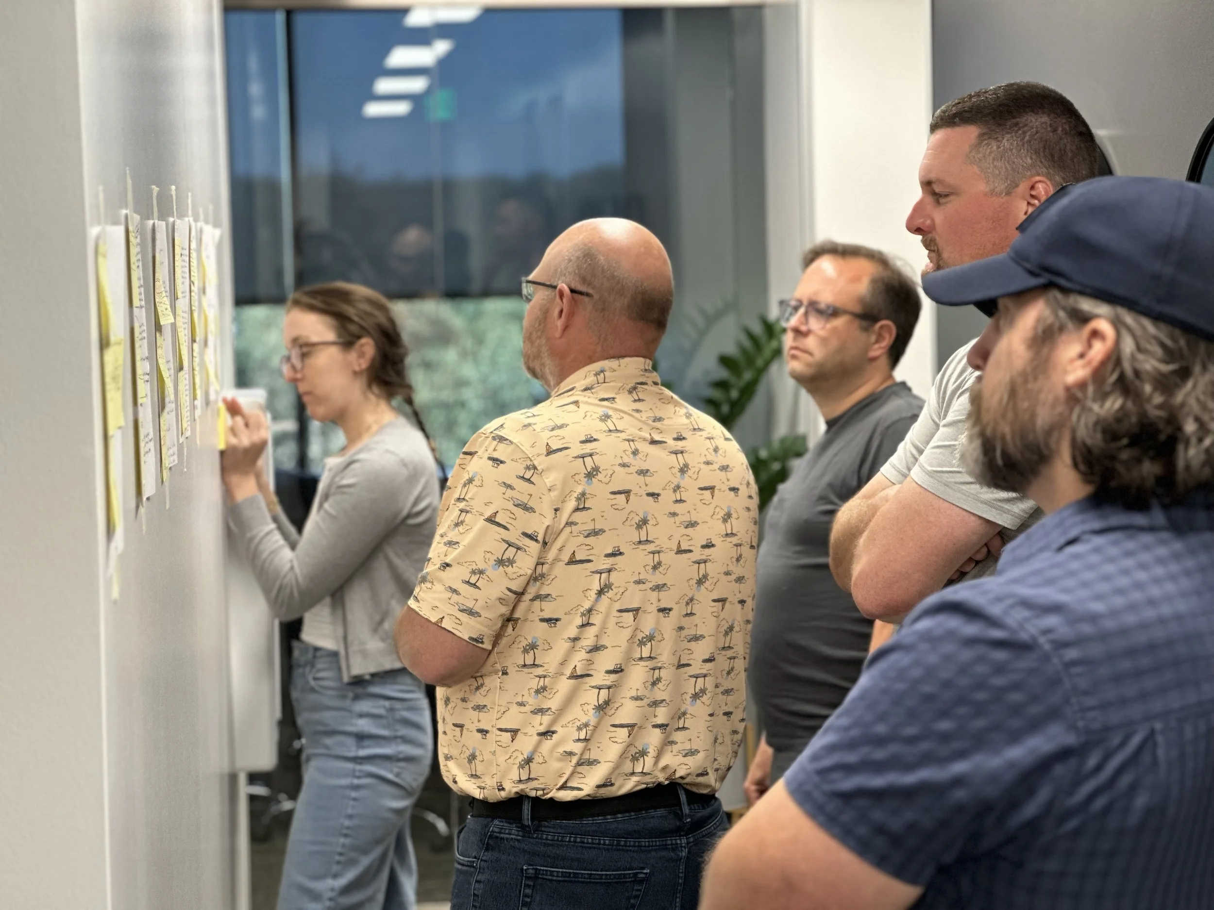 Five people in a meeting room, looking at notes on a wall, engaging in a discussion or brainstorming session.