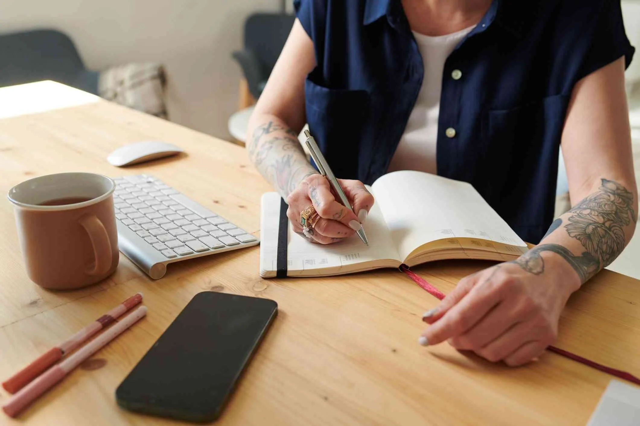 Person with tattoos writing in a planner at a wooden desk with a coffee mug, computer keyboard, smartphone, and pens.