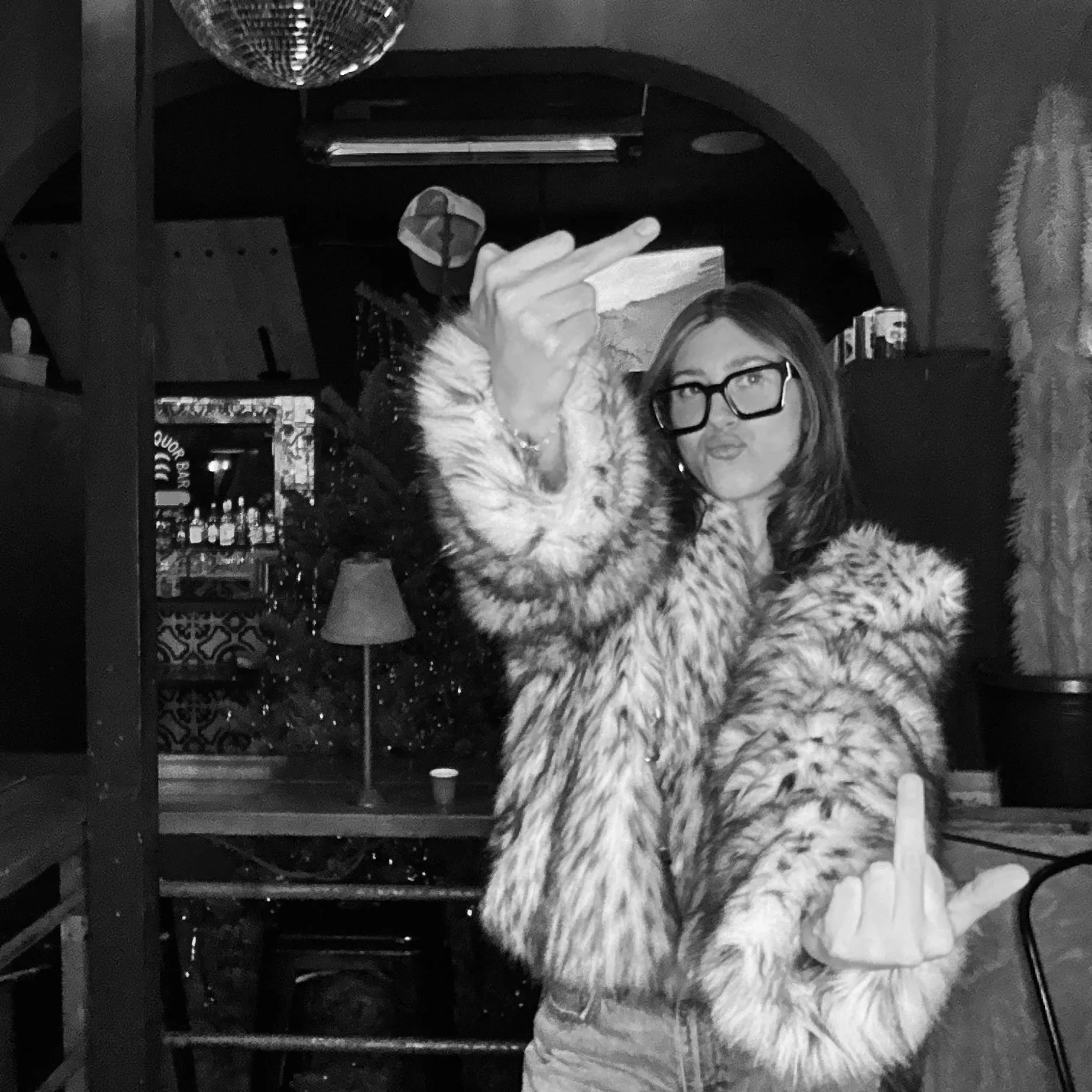 Person in faux fur coat and glasses making rude gestures in a dimly lit room with disco ball.