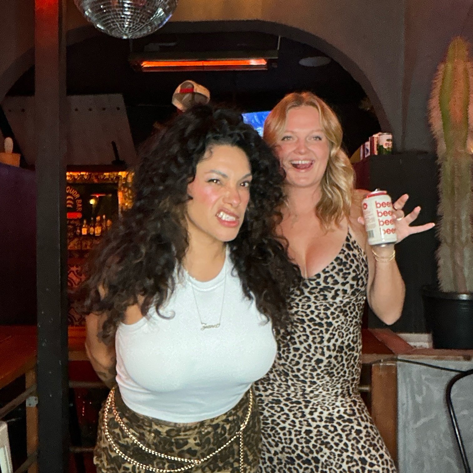 Two women posing in a bar, one wearing a white tank and leopard-print skirt, the other in a leopard-print dress, holding a beer can. There is a disco ball above and a large cactus in the background.
