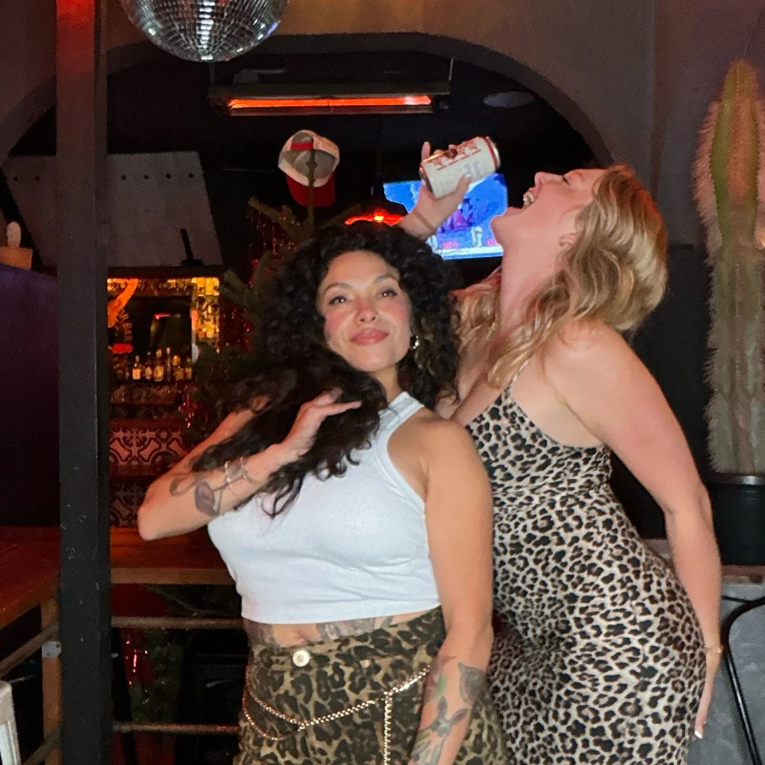 Two women posing playfully at a bar, one drinking from a can, wearing leopard print outfits.