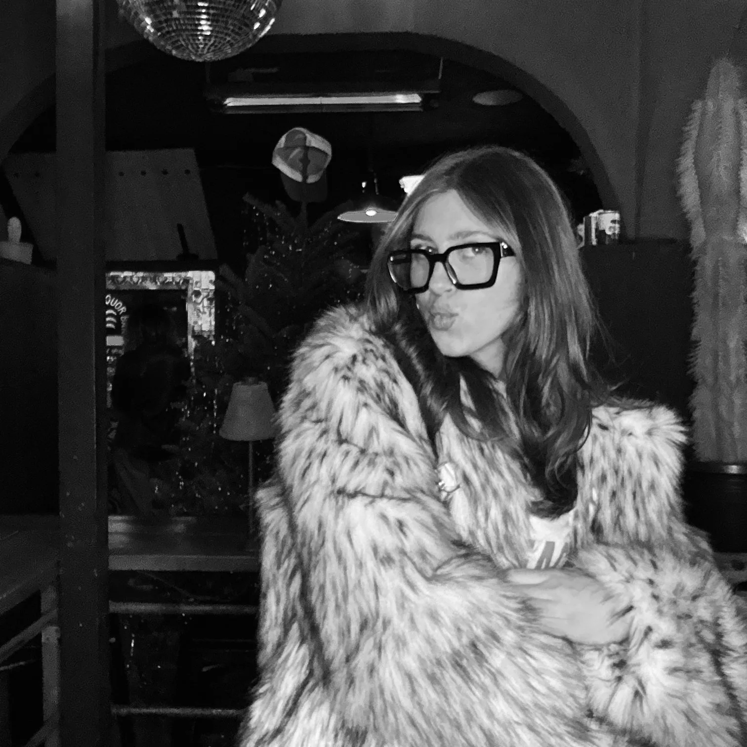 Black and white photo of a person wearing large glasses and a faux fur coat, posing indoors with a playful expression. A disco ball and ambient lighting are visible in the background.