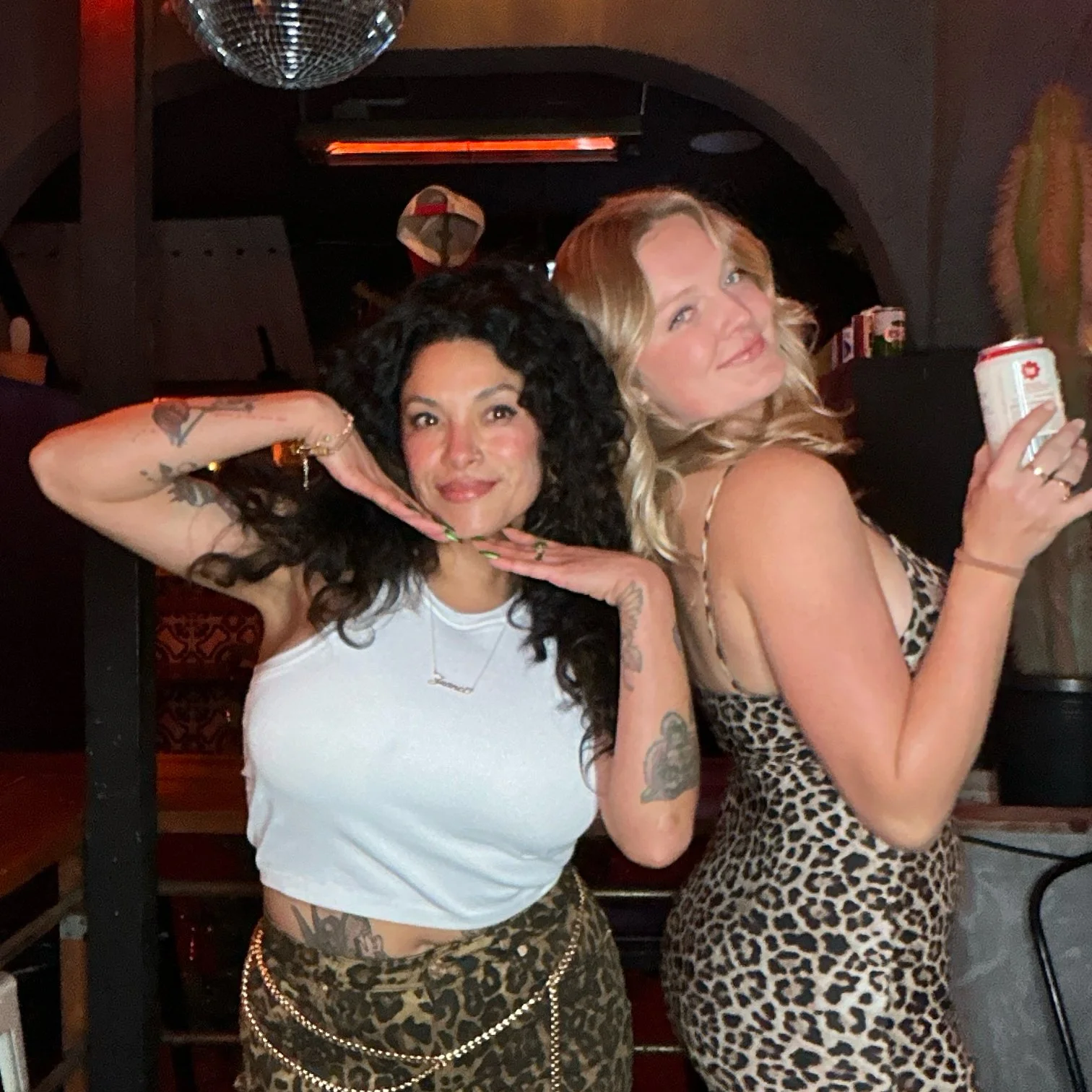 Two women posing in a bar, one with curly hair and a white top, the other with wavy hair, leopard print dress, holding a drink.