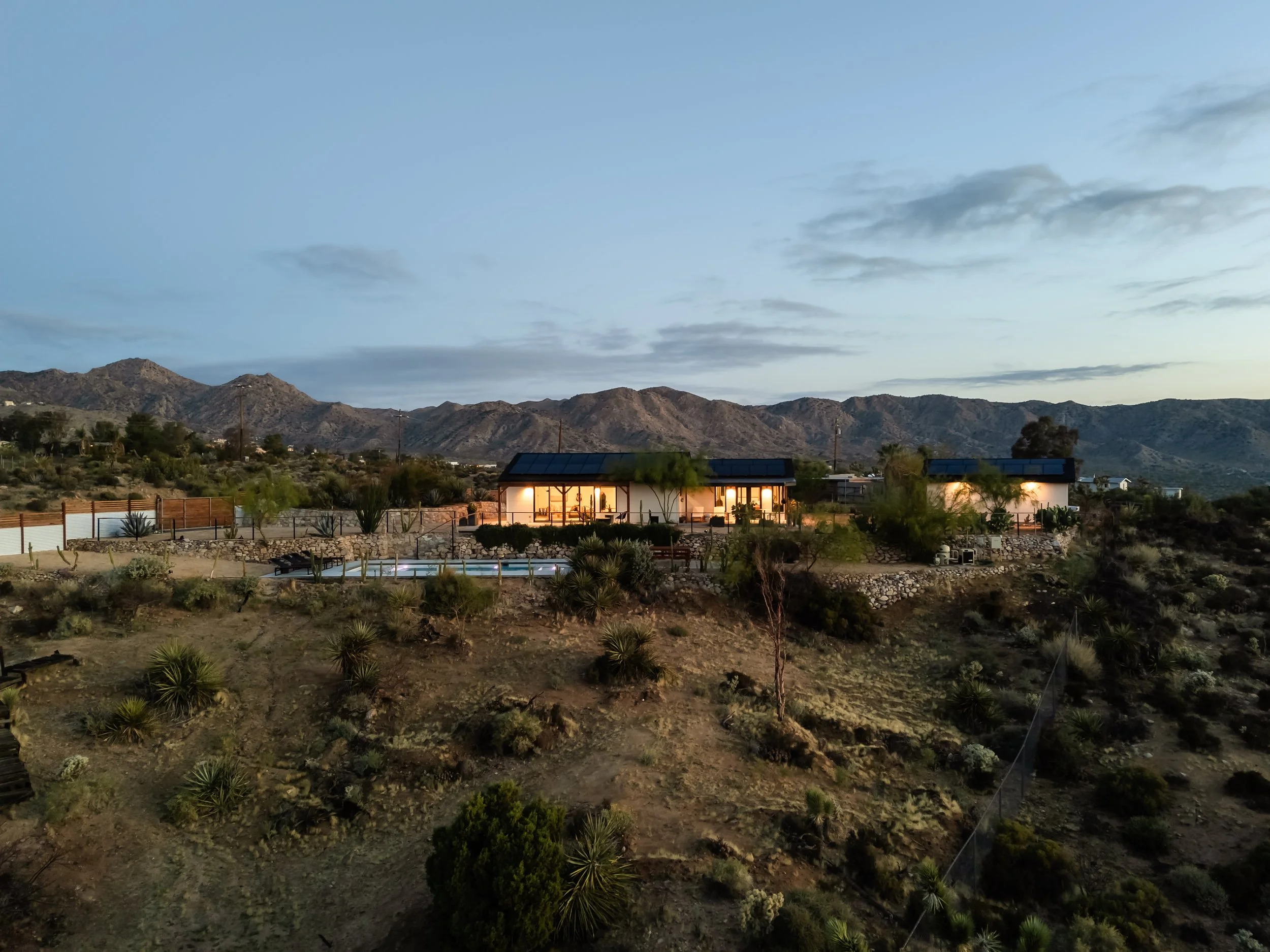 A house with lights on, situated on a hillside with desert vegetation, mountains in the background, and a cloudy sky at dusk.