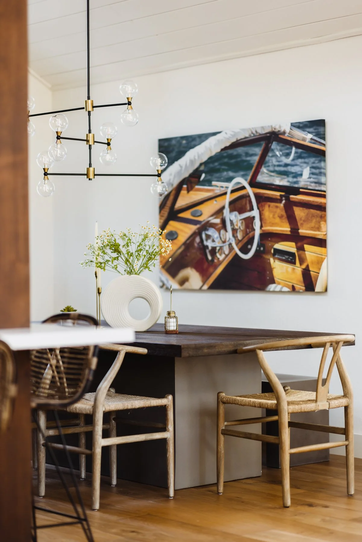 Interior of a dining room showing a wooden table with decorative items, wooden chairs, a modern chandelier, and a large piece of artwork on the wall depicting a boat's interior.
