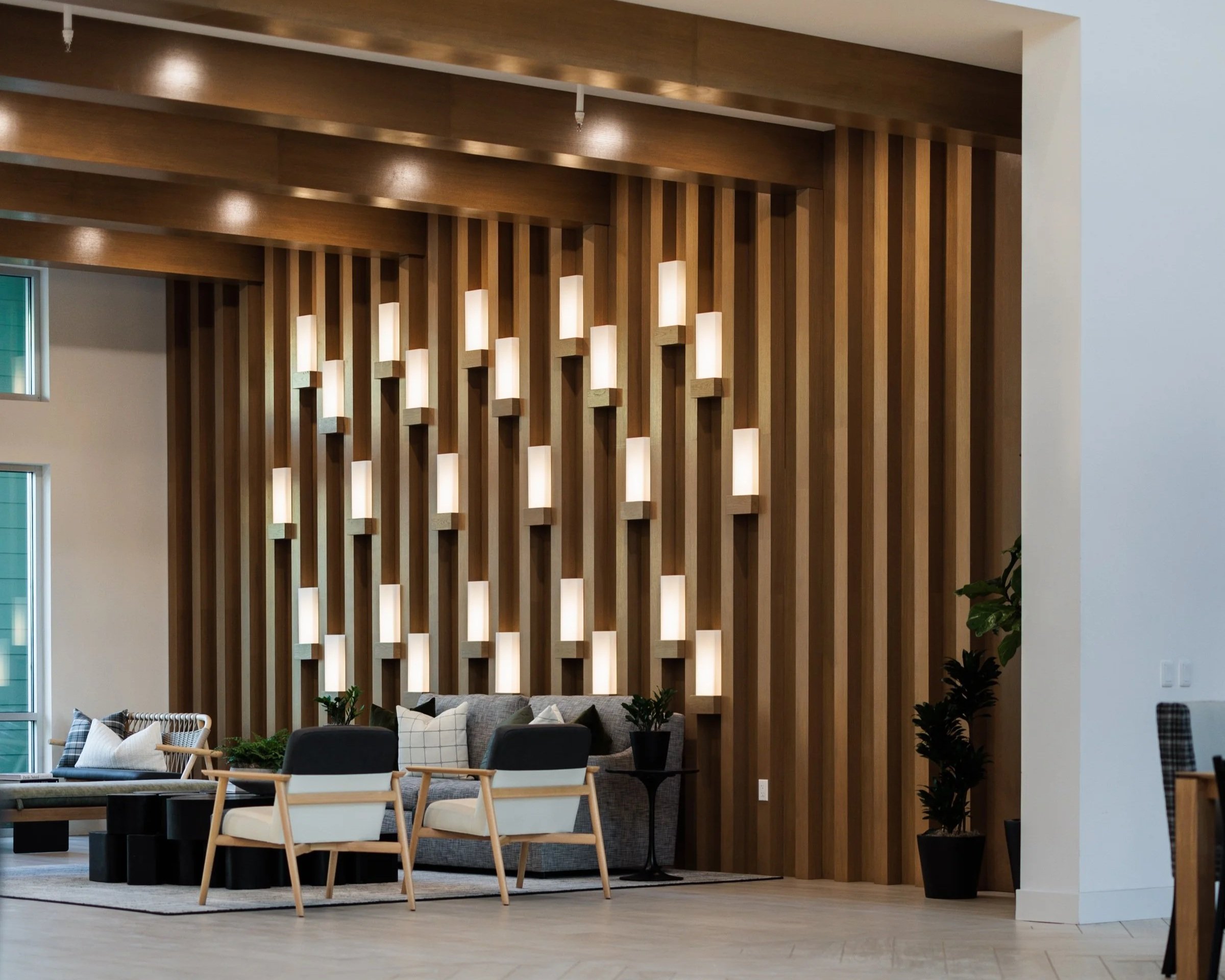 Modern interior of a living room with wooden wall panels, decorative lighting, a gray sofa, white and black chairs, a coffee table, and green houseplants.