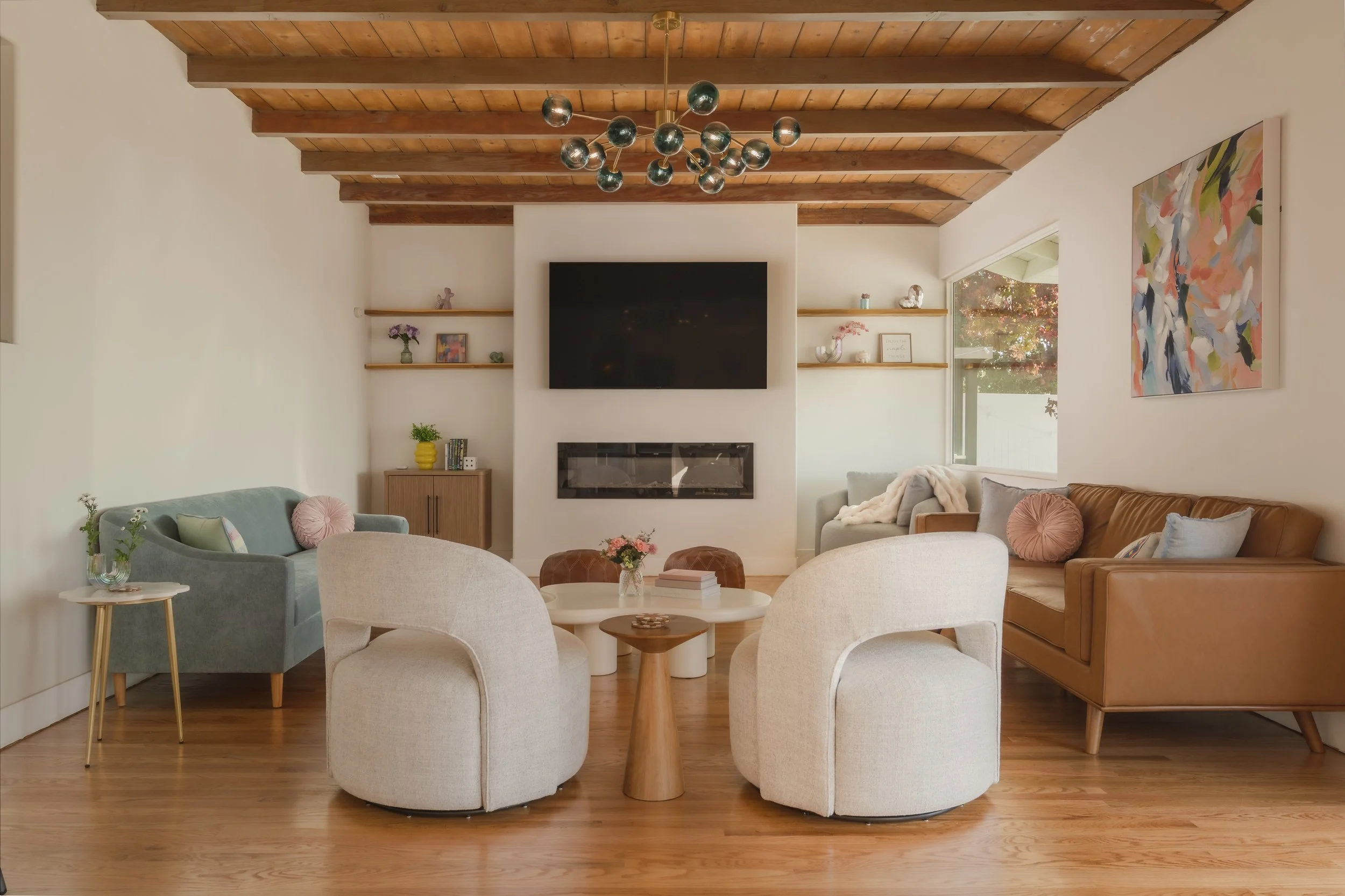 Living room with wooden ceiling, modern furniture including a gray armchair, a tan sofa with pillows, white armchairs, a small side table, a round coffee table with flowers and books, wall-mounted TV, shelves with decor, and a window with outdoor vie