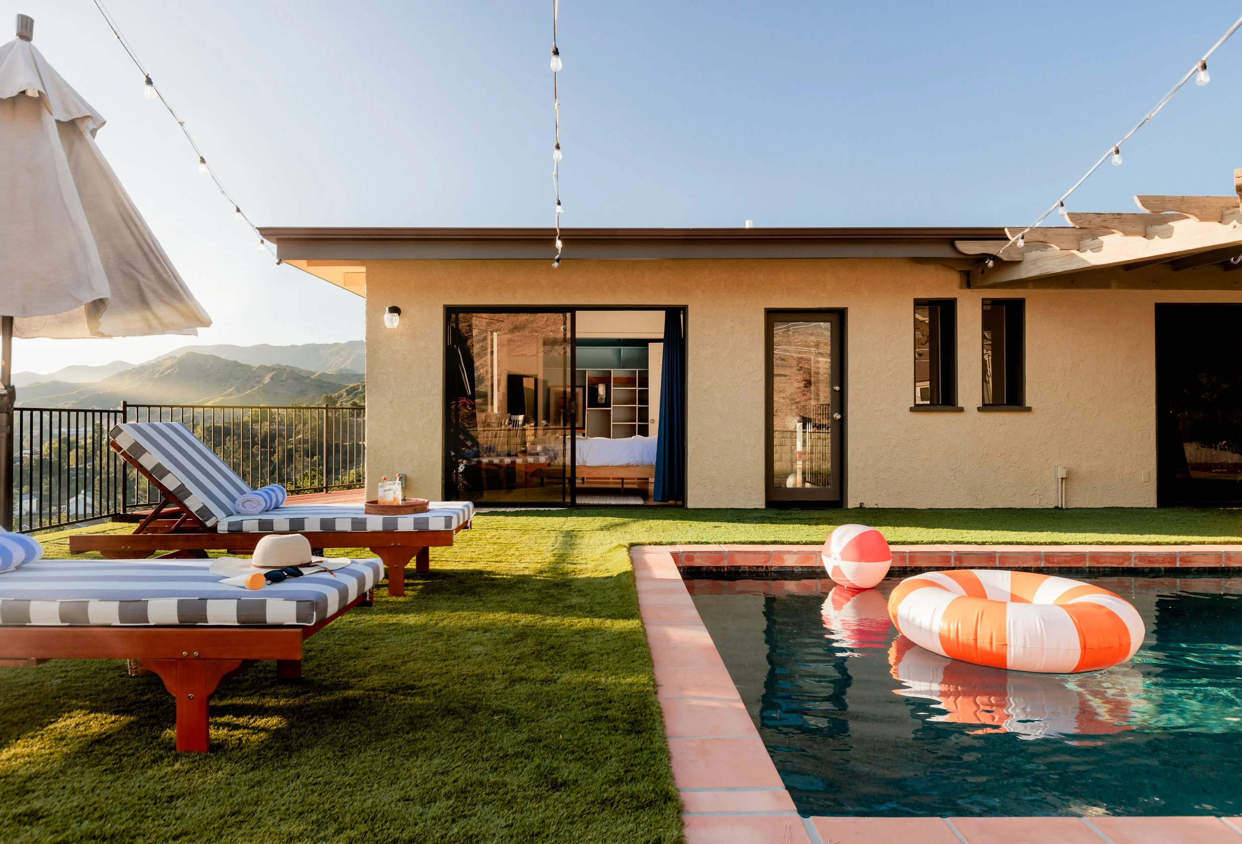 A backyard with a swimming pool featuring orange and white floaties, surrounded by a grassy area with striped lounge chairs, a large umbrella, and a view of mountains in the background. There are string lights hanging above, and a house with sliding 