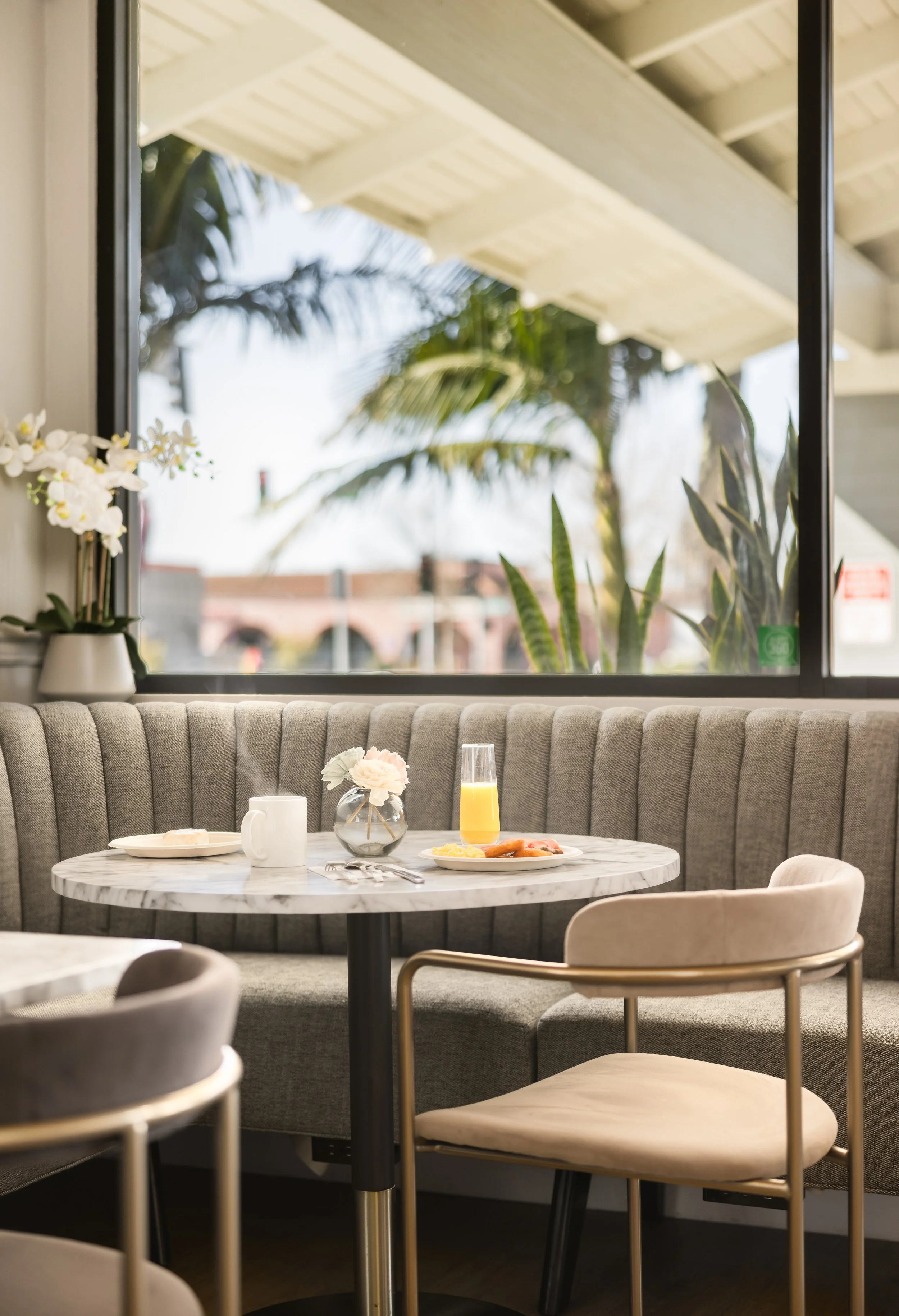 A breakfast table with a marble top set for one, including a plate of food and a glass of orange juice, in a cozy restaurant corner with a large window showing outdoor greenery.