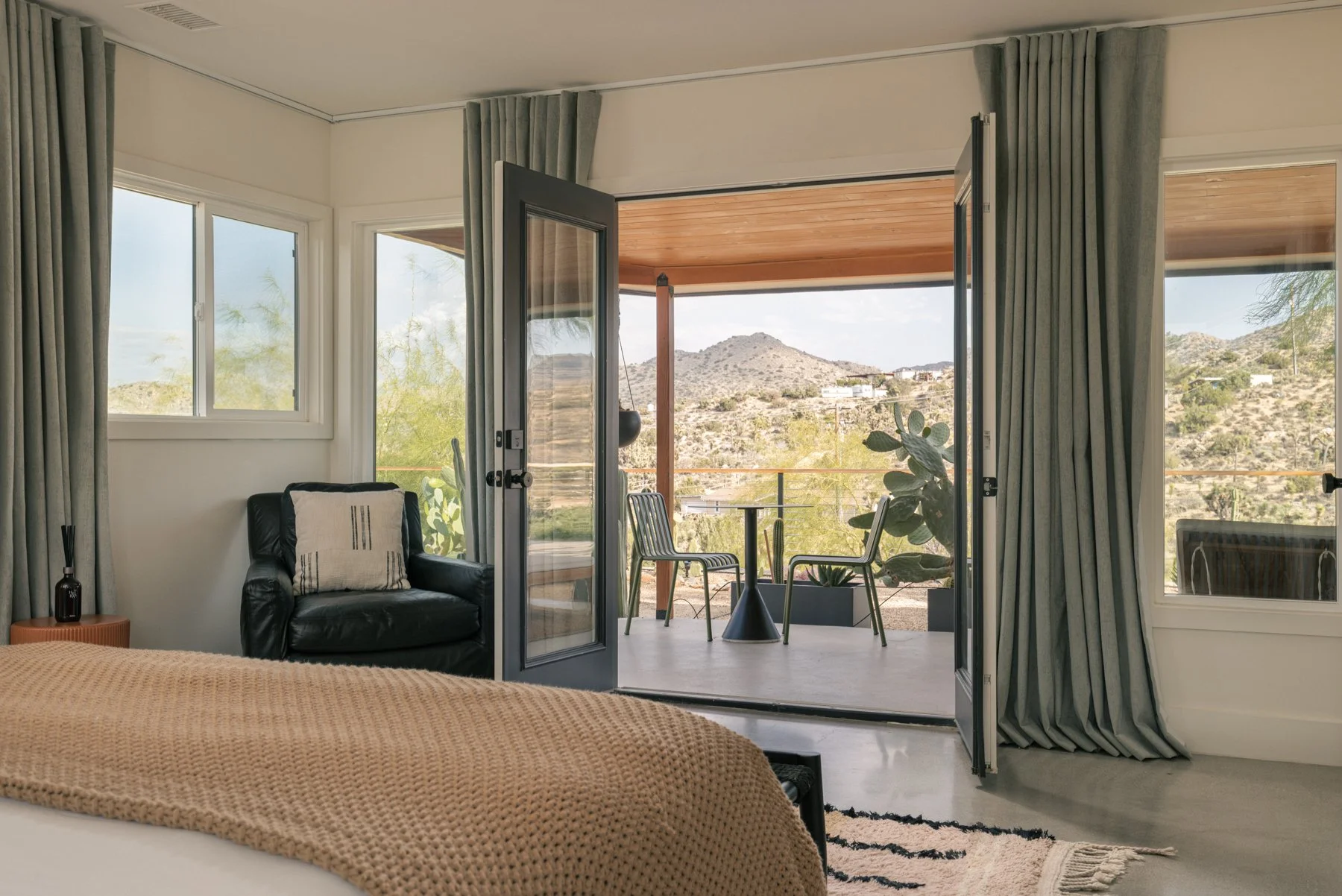 A bedroom with an open door leading to a balcony, showing a mountain landscape in the background. The room has a bed, a black leather armchair with a cushion, large windows with curtains, and a rug on the floor.