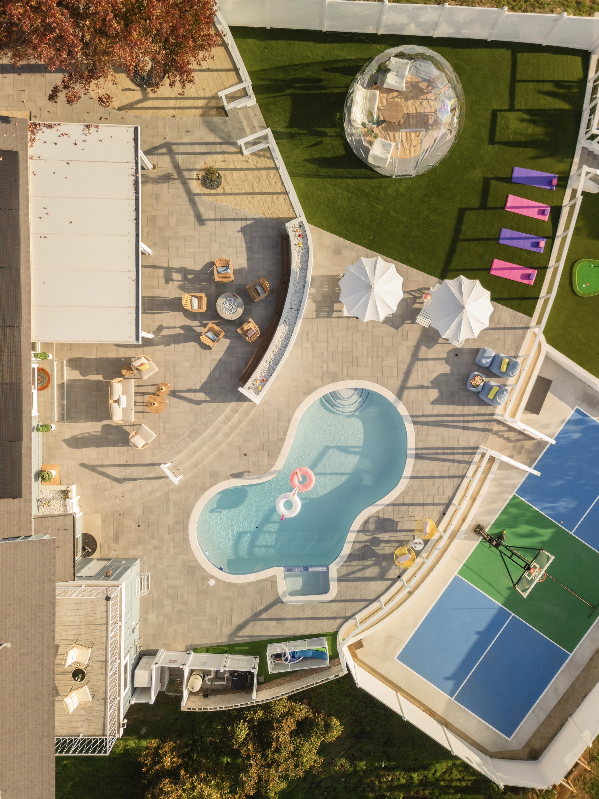 Aerial view of a backyard with a swimming pool, outdoor seating area, a sport court, and a lush green lawn with a transparent dome-shaped structure.
