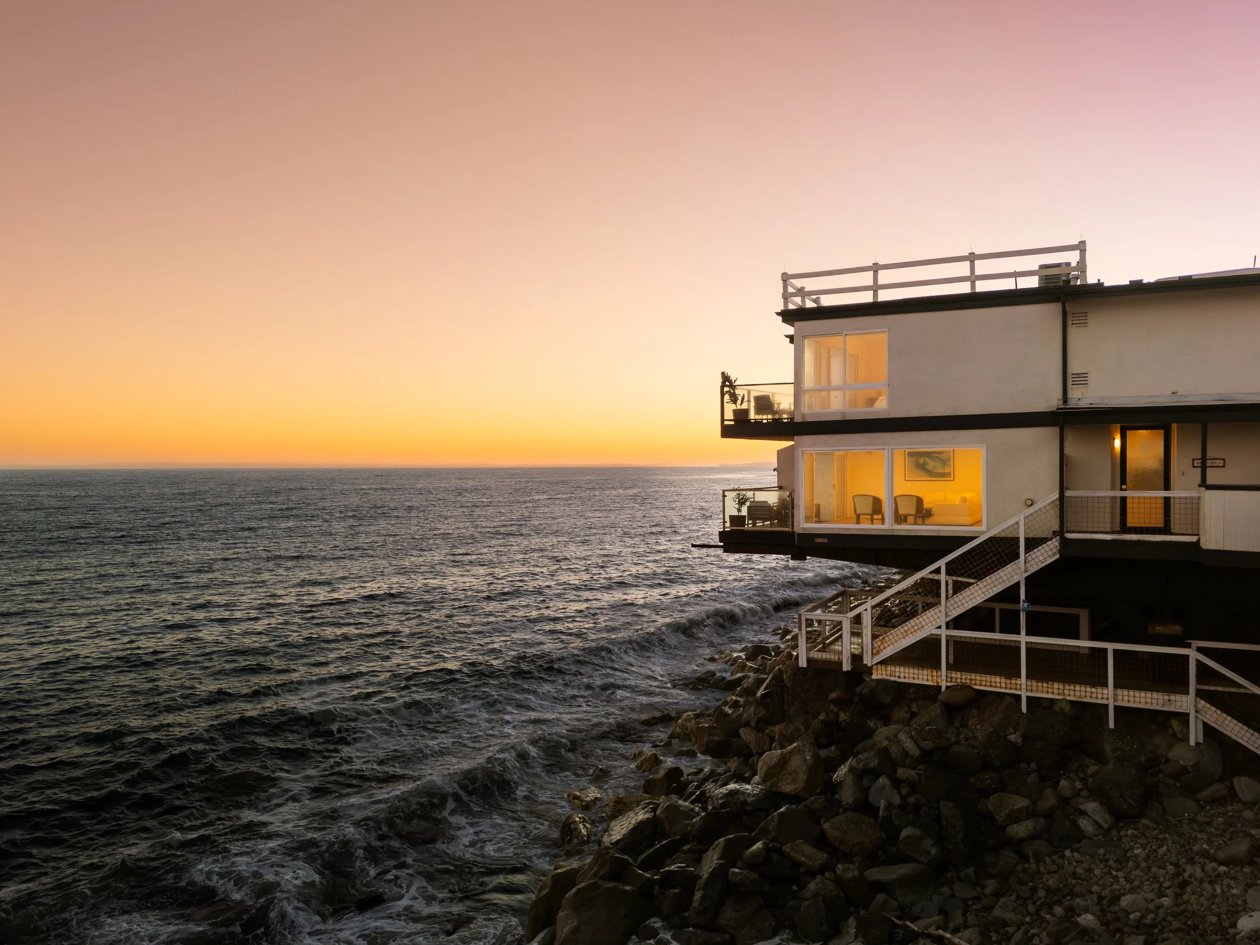 A beachfront house with large windows illuminated from inside, built on rocks by the ocean at sunset.