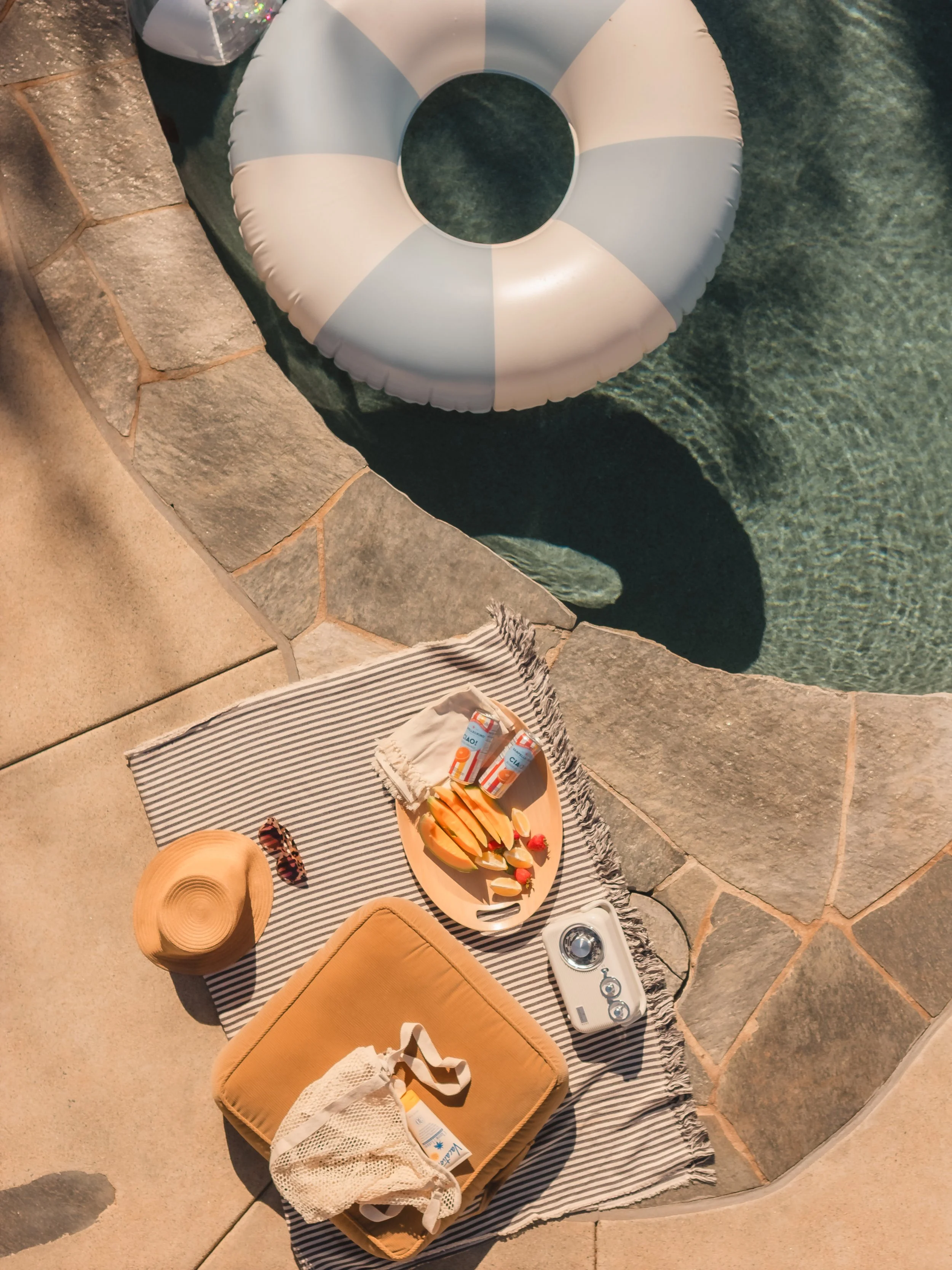 Picnic setup by a swimming pool featuring a striped blanket, a beige cushion, a straw hat, a pack of sunscreen, a tray with sliced fruits, and a small camera, with a life preserver floating in the pool nearby.