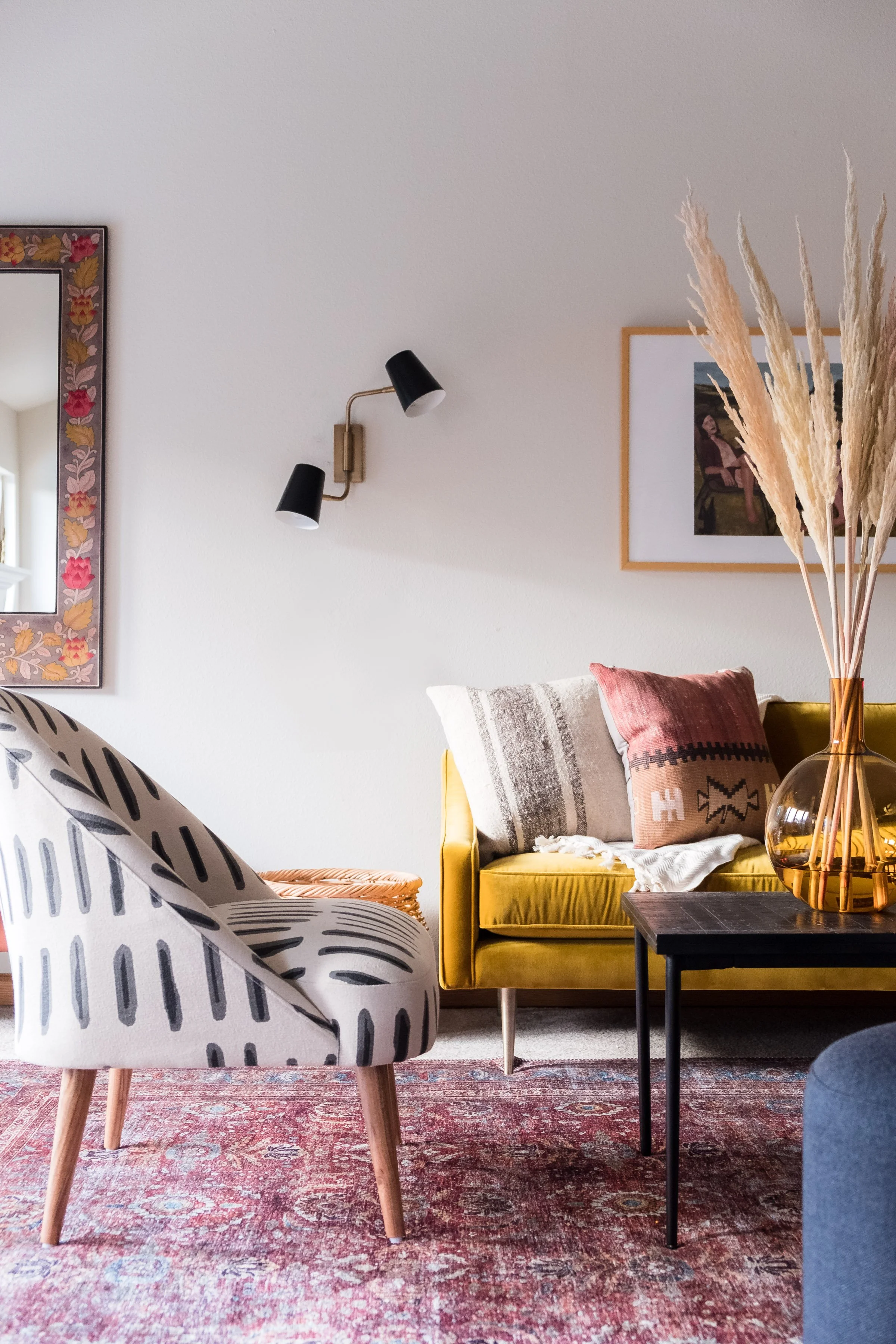 A living room with a patterned armchair, yellow sofa with cushions, a black coffee table with a copper vase of pampas grass, wall art, and a pink patterned rug.