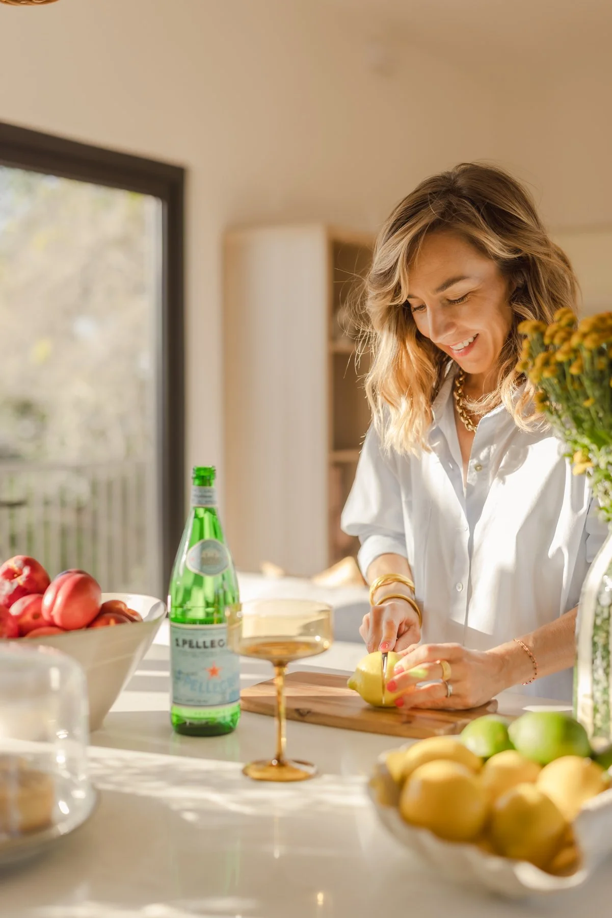 A woman chopping a yellow apple in a brightly lit kitchen with fresh apples and lemons on the counter, a bottle of sparkling water, and a champagne glass nearby.