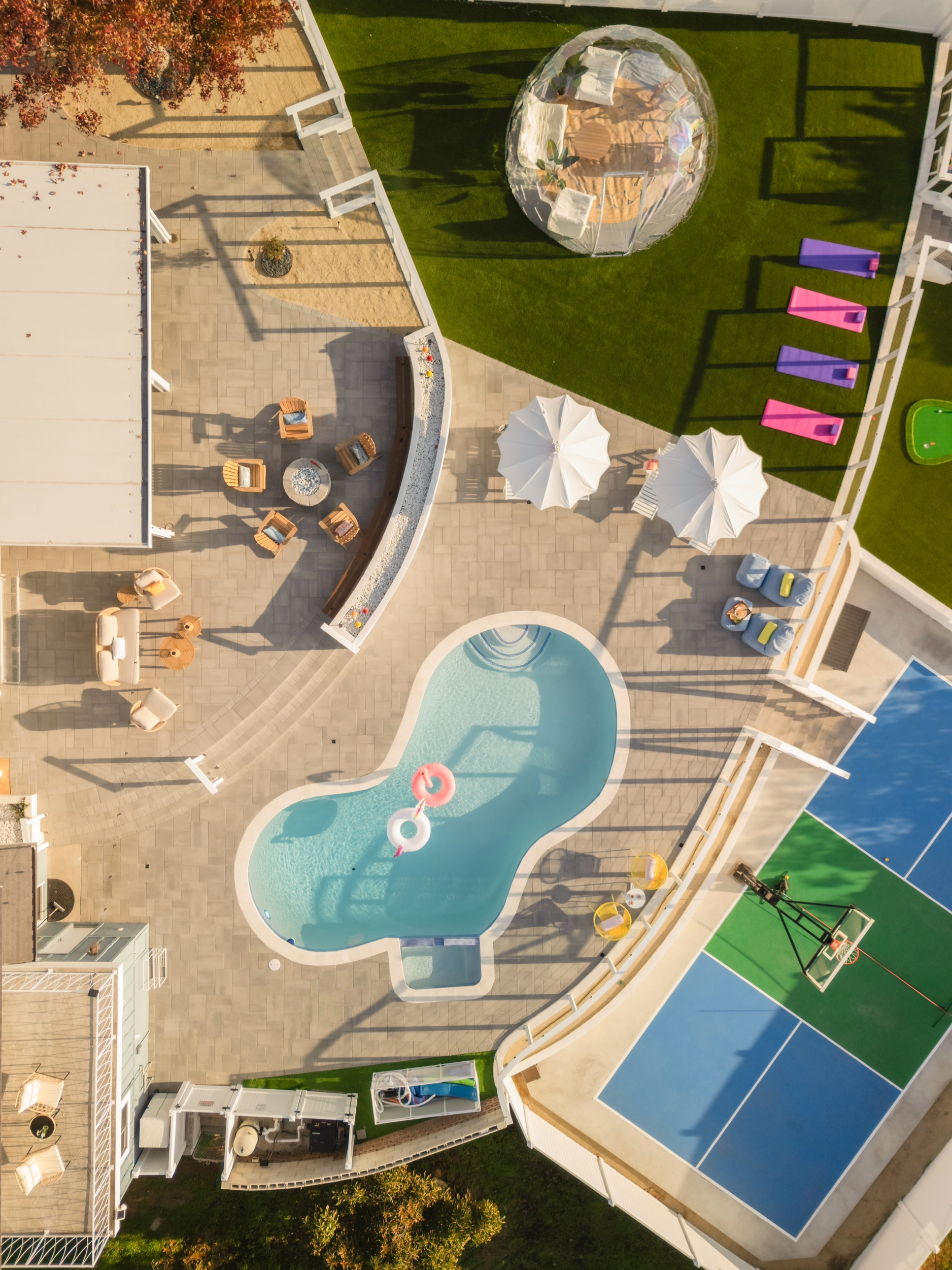Aerial view of a backyard with a swimming pool, patio seating, pink and purple lounge chairs, a trampoline, a basketball hoop, and a covered seating area with umbrellas.