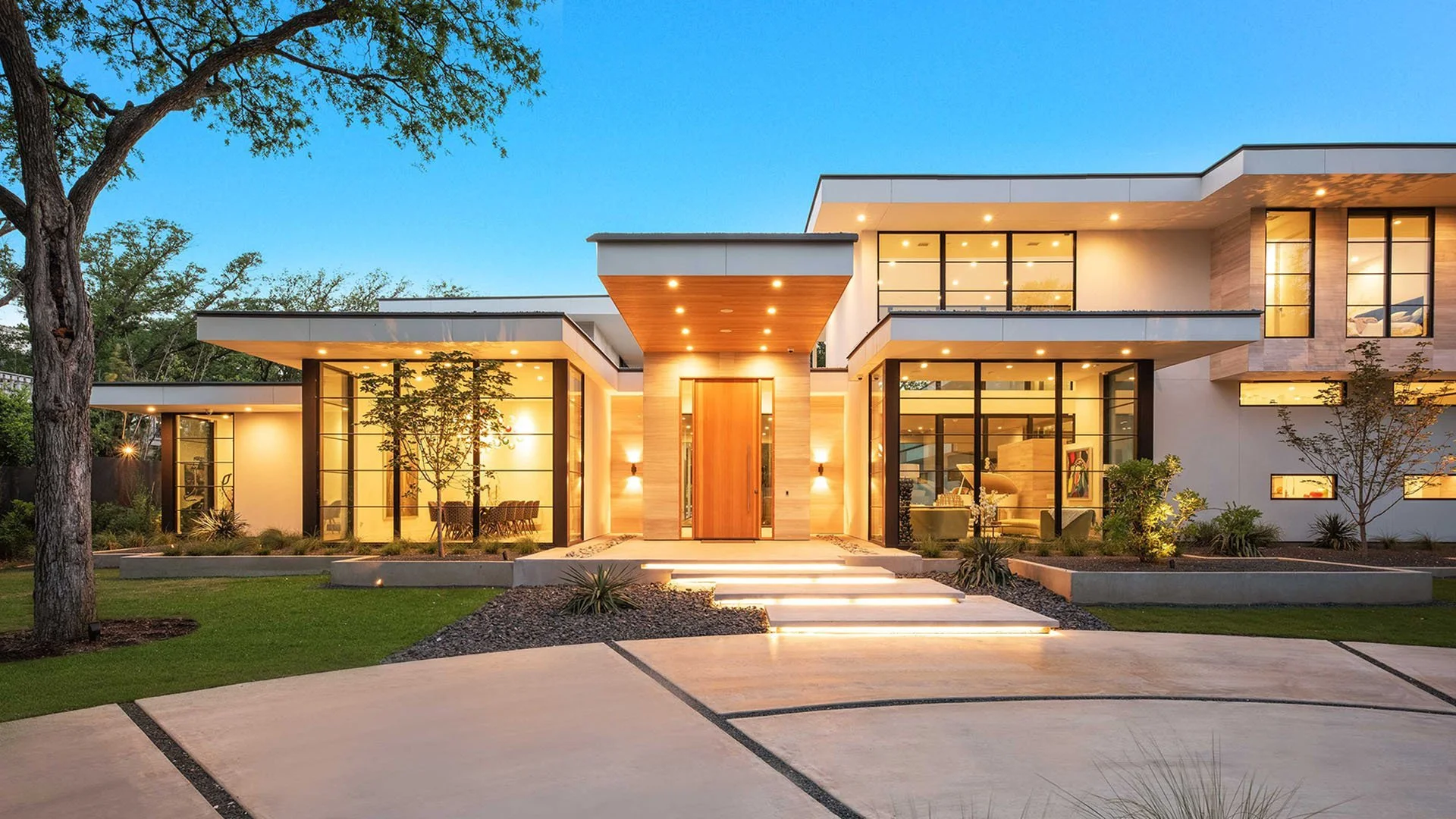 DARGO Construction - Modern two-story house with large glass windows, illuminated exterior lighting, a staircase leading to the front door, landscaped yard with trees, green grass, and a concrete driveway.