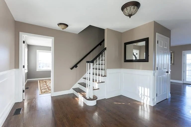 Interior view of a home's hallway with beige walls, white wainscoting, and hardwood floors. A staircase with white balusters and a black handrail leads upstairs. There's a mirror on the wall next to a closed white door. An open door reveals a room with a window and a rug. Natural light fills the space.