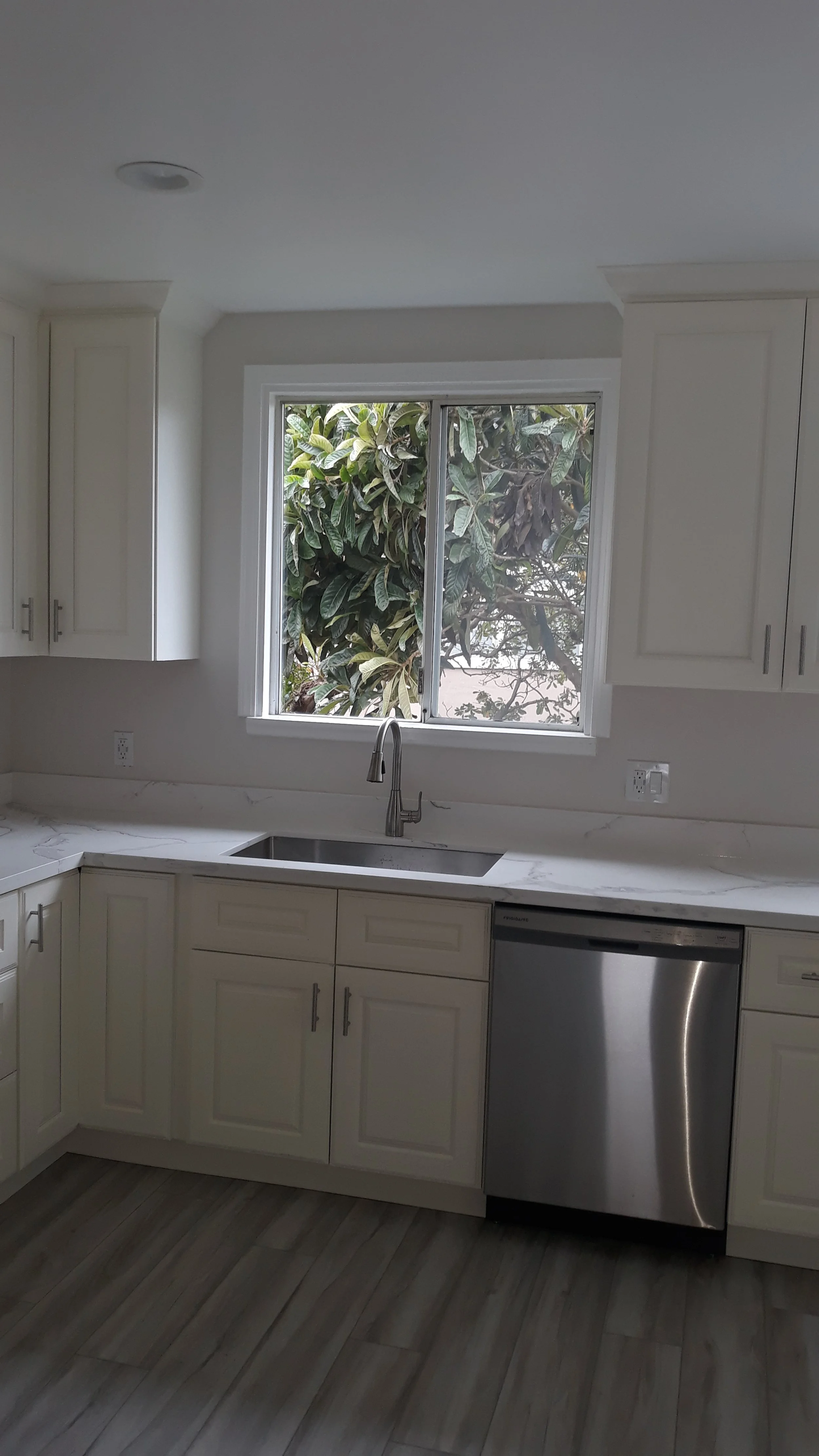 DARGO Construction - A modern kitchen with white cabinets, a marble countertop, a stainless steel dishwasher, and a window overlooking leafy trees.
