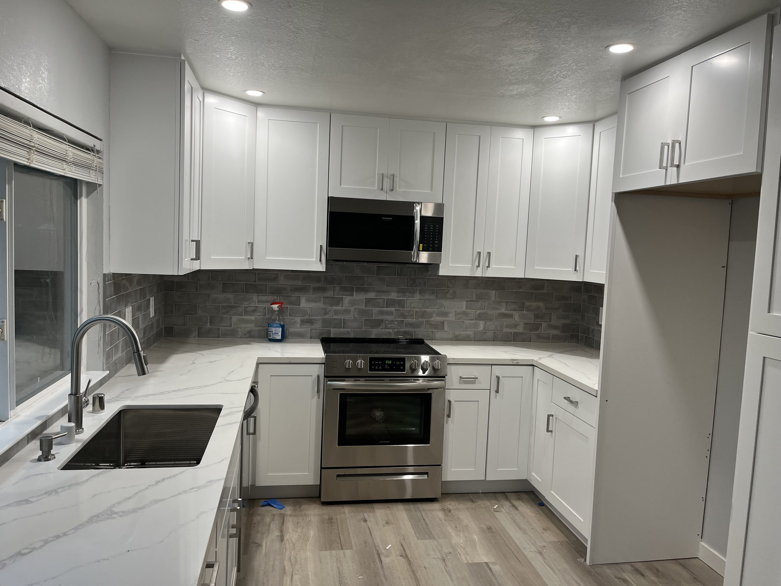 Modern kitchen with white cabinets, gray backsplash tiles, stainless steel oven and microwave, marble countertop, and a window with blinds.