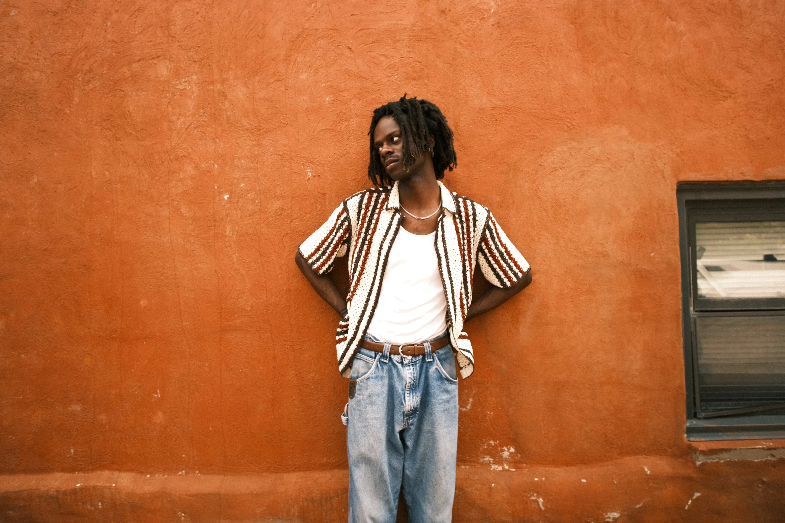 A young man with dreadlocks wears a patterned short-sleeve shirt, white t-shirt, and light-wash jeans, leaning against an orange wall with hands behind his back.