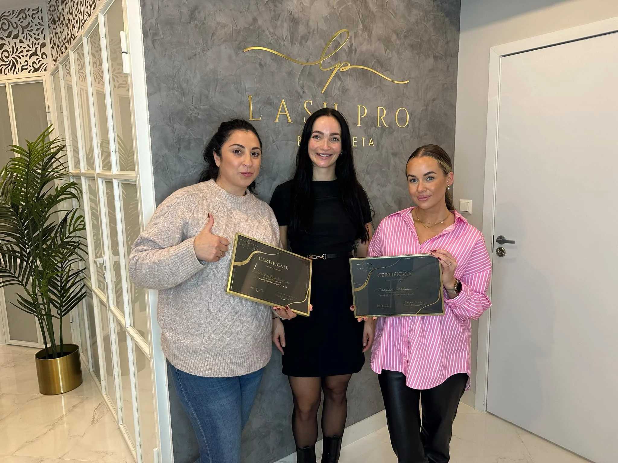 Tre kvinner står foran en vegg med logoen til laspro og holder opp sertifikater, alle smilende og festlige, i et rom med delvis glassdører og en plante.