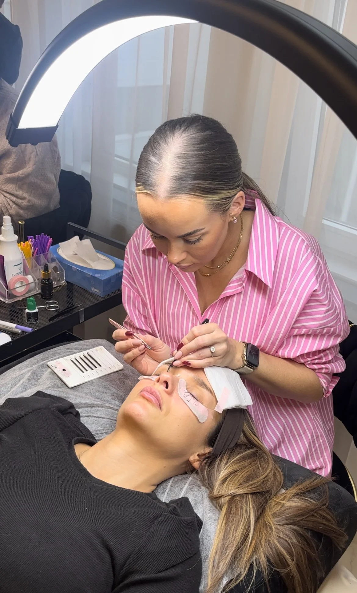 En kosmetolog utfører en øyenlokkforbedring på en kvinne som ligger ned, mens en annen kvinne holder henne rolig på hodet.