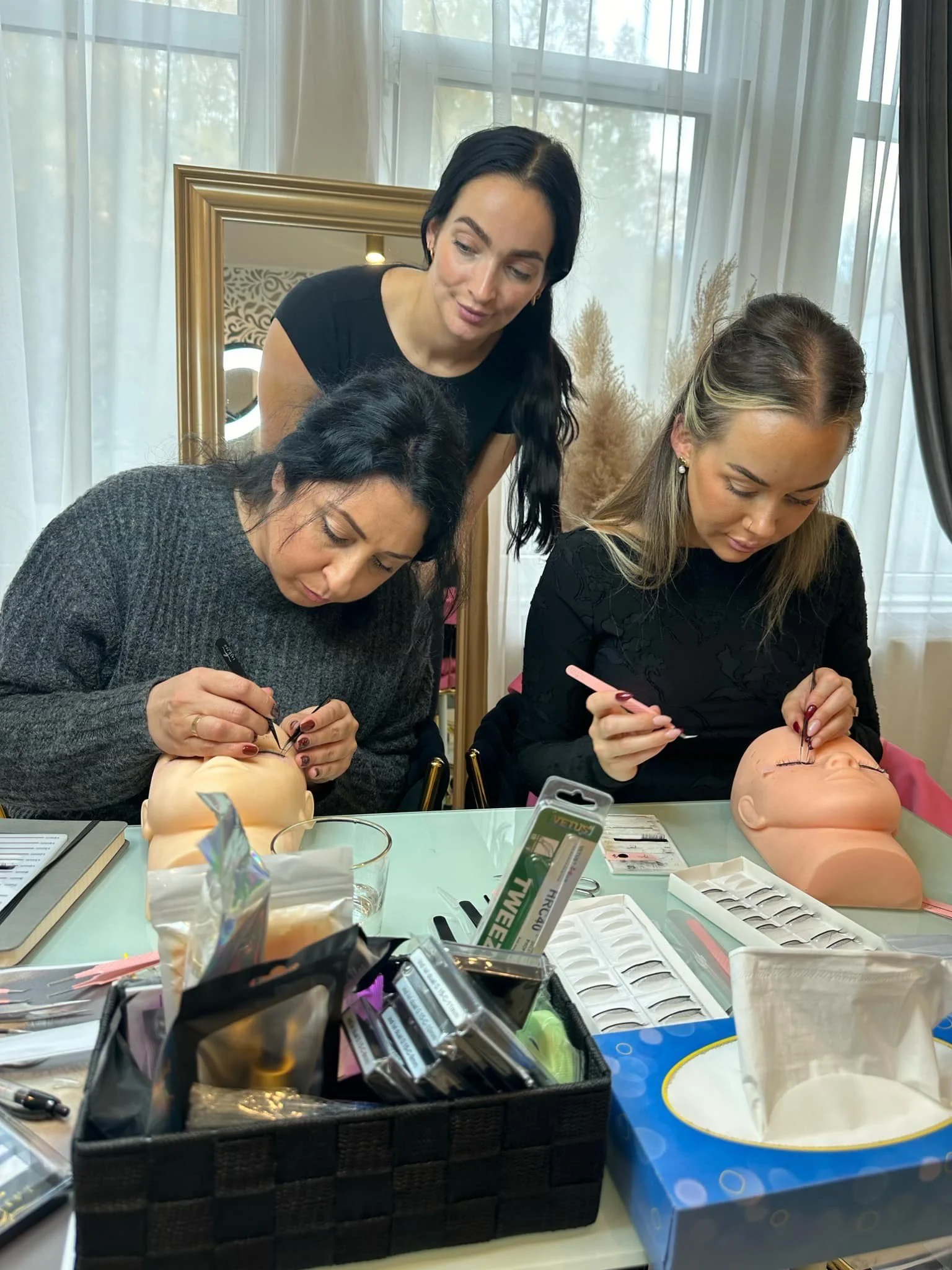 Tre kvinner trener på å sette falske øyevipper på mannequiner med øyenlokk i en innendørs setting. To av kvinnene jobber intens med å feste vippene, mens den tredje står bak dem og observerer.