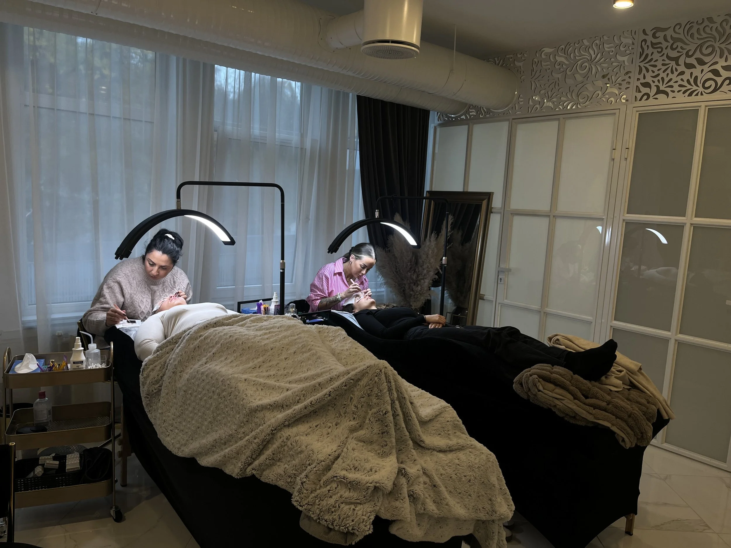 To personer får behandling på skjønnhetssalong, hver på sin seng, under salongbelysning, med klær og峒er på, i et rom med store vinduer og blonder på gardinene.