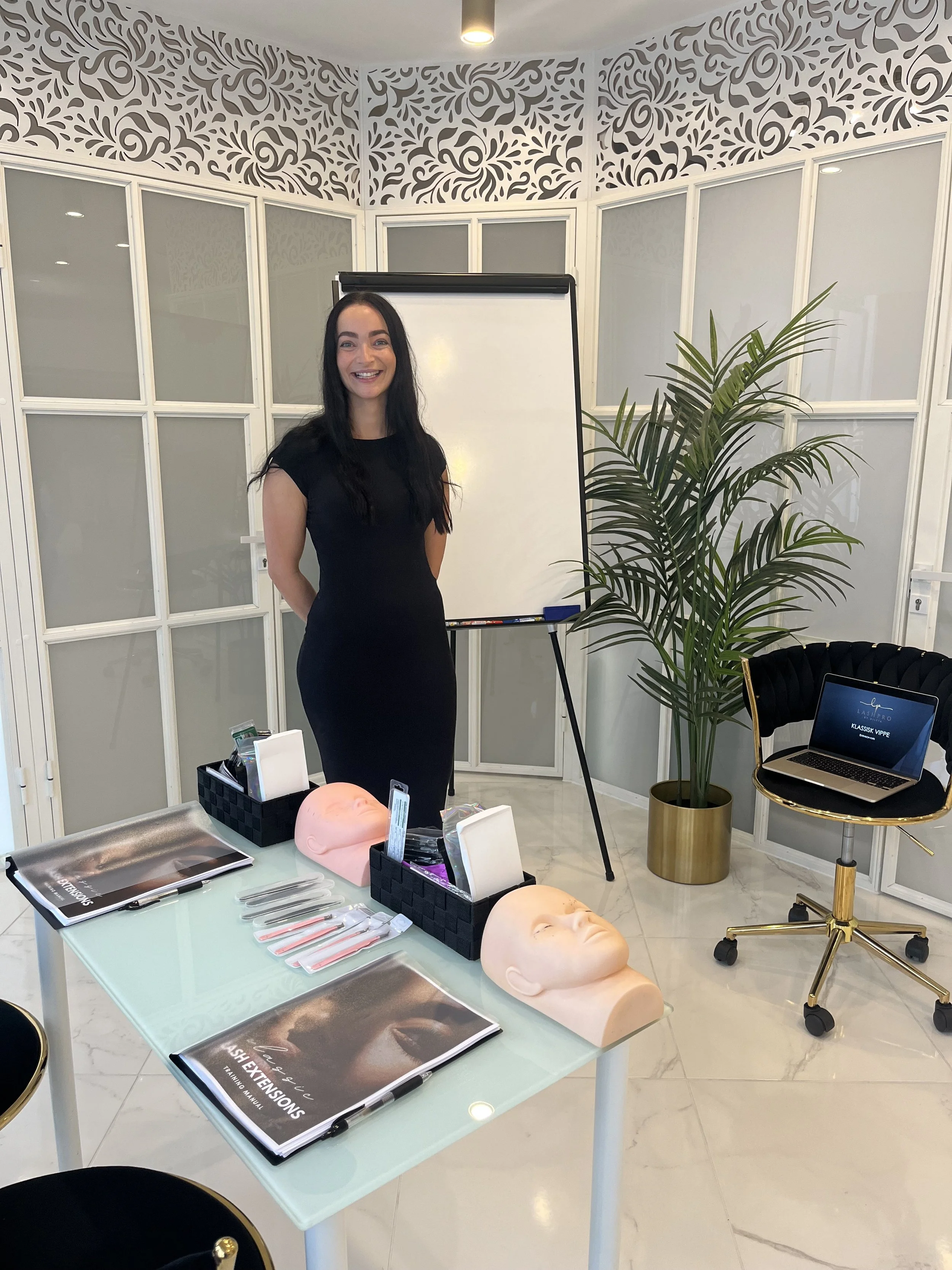 En kvinnelig instruktør står foran en whiteboard i et klasserom, med medisinske modeller av ansikter og redskaper på et bord foran henne.