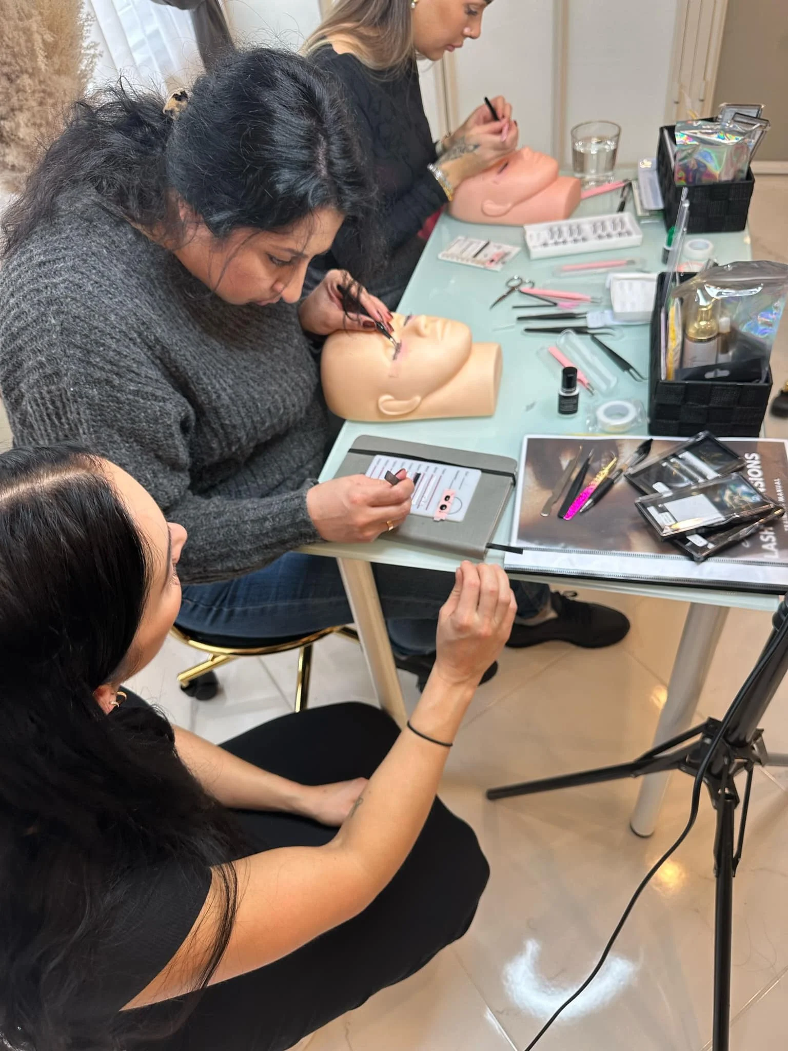 To personer som jobber med å påføre makeup på mannequinhoder i et rom med bord og ulike skjønnhetsverktøy og produkter.