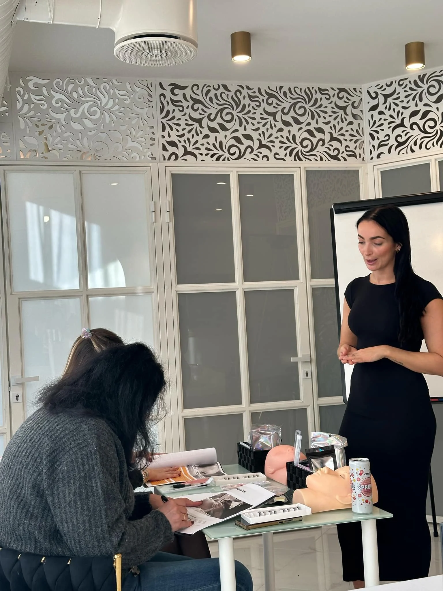 En kvinne holder et foredrag foran en gruppe mennesker i et rom med dekorative veggpaneler, bord med medisinske modeller og utstyr, og personer som ser ut til å ta notater.