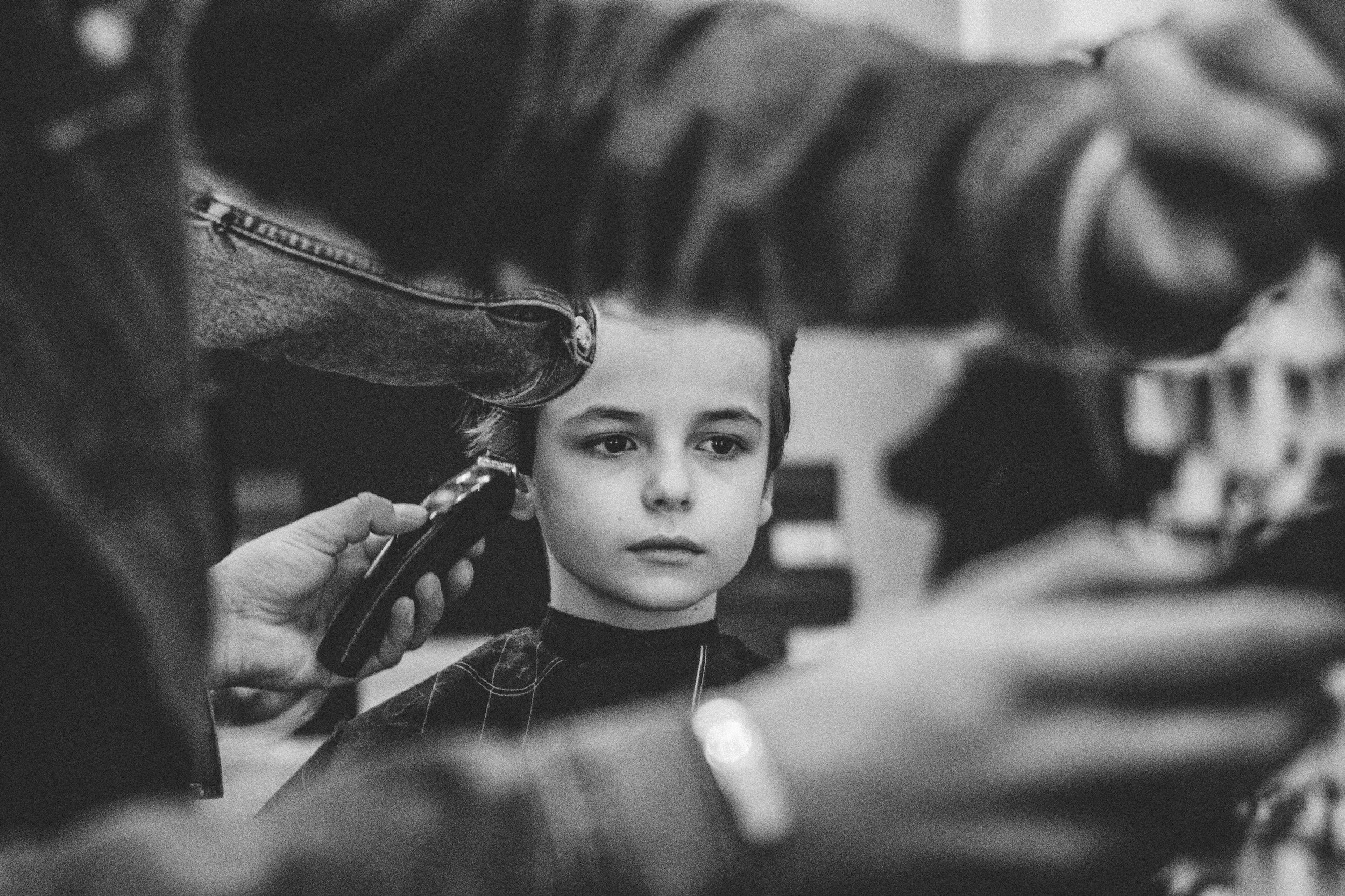 A young boy getting a haircut in a barber shop, sitting in a chair while a barber trims his hair. The scene is in black and white.