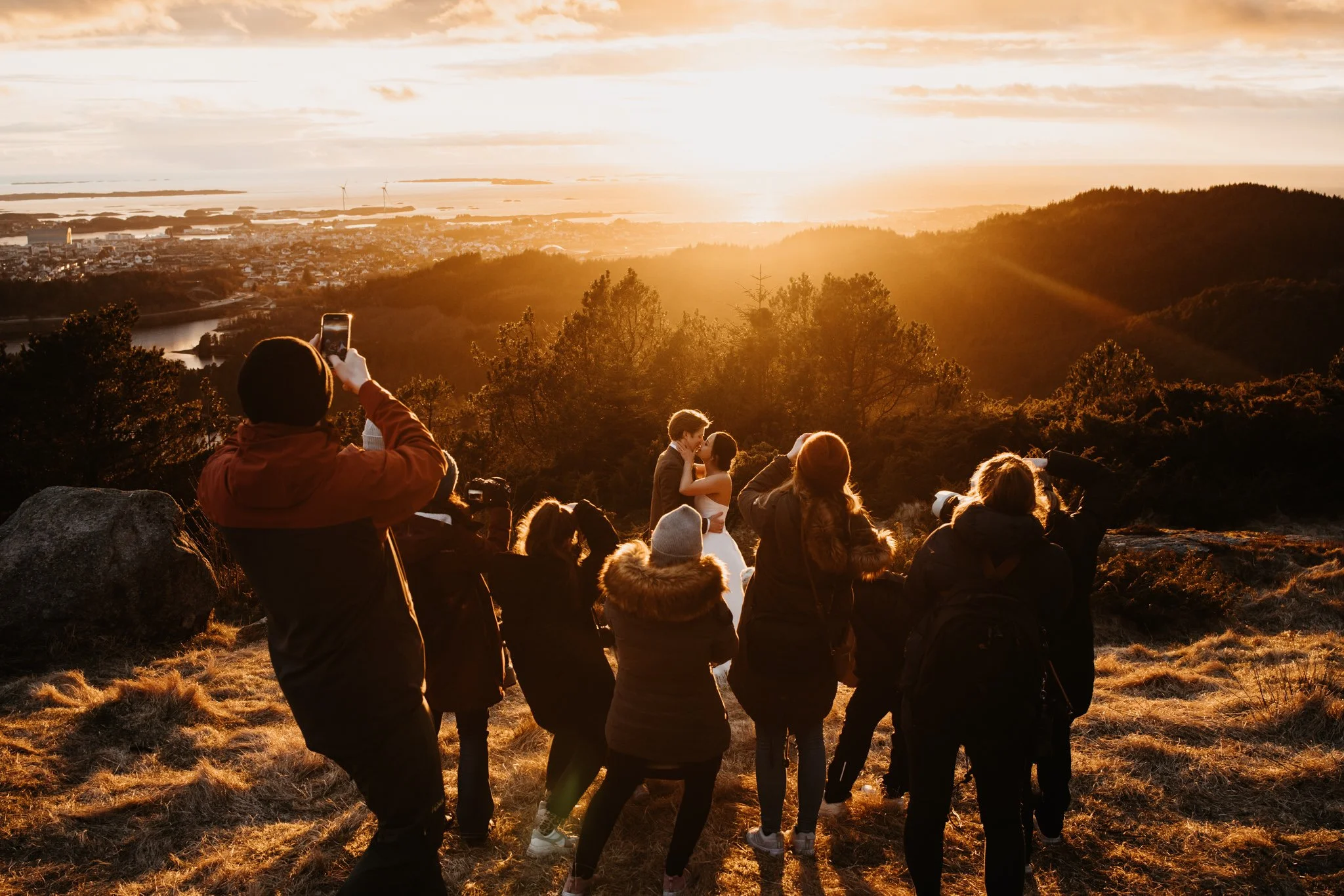 Et bryllupsbilde av et par som kysser under en solnedgang, omgitt av fotografer og tilskuere på en åsside.
