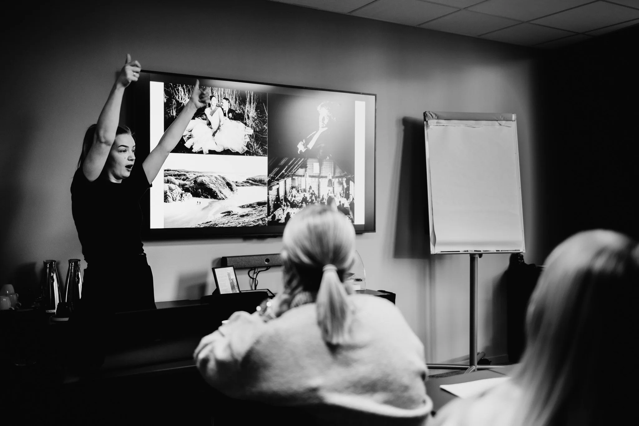 En kvinne holder en presentasjon foran en skjerm med bilder, mens to personer ser på og lytter.