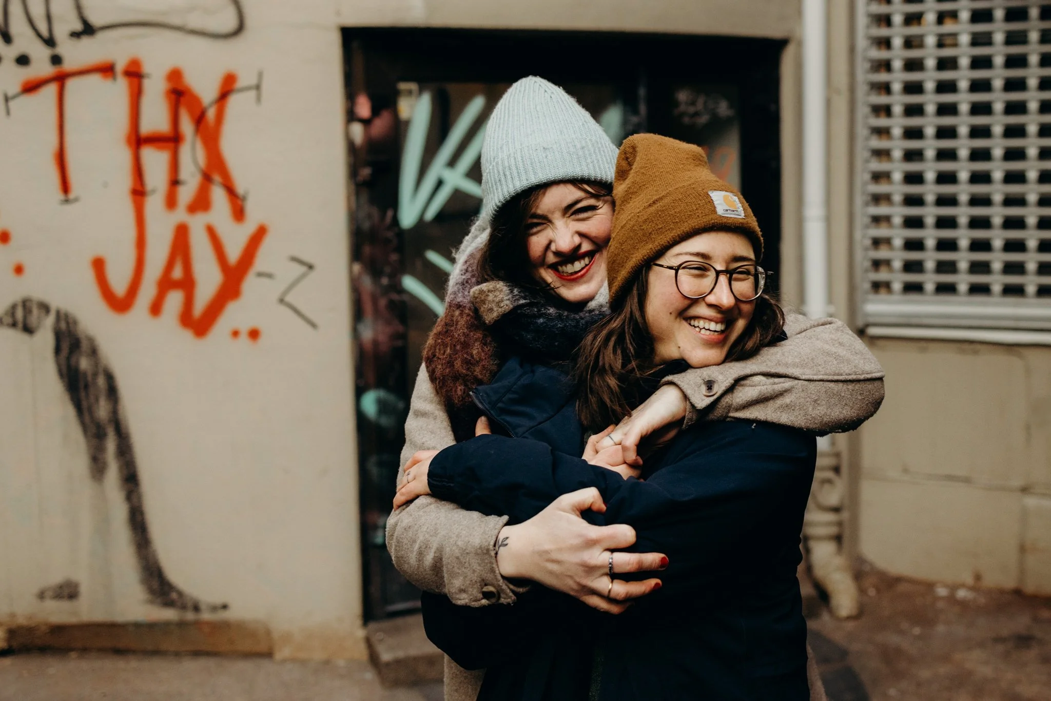 To kvinner som smiler og klemmer hverandre, iført varme klær og luer, utendørs i en gate med graffiti og lys i bakgrunnen.