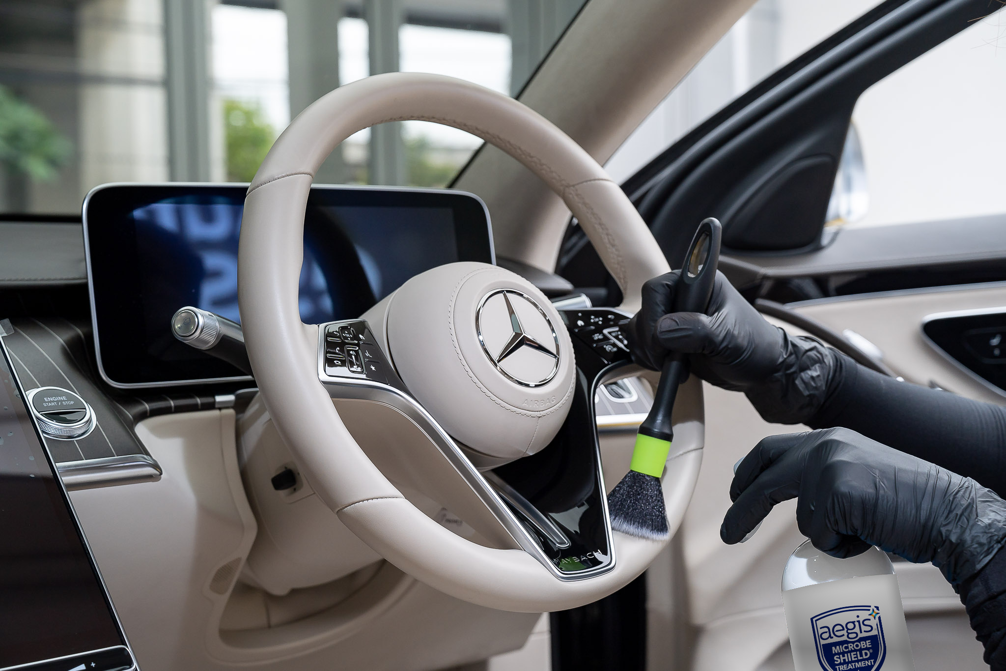 Close-up of a person's hand wearing a black glove, pressing a button on a white Mercedes-Benz steering wheel inside a car. The dashboard display is turned off, and a cleaning product labeled 'Aegis Microbe Shield' is visible in the foreground. The interior is modern with digital controls and a sleek design.
