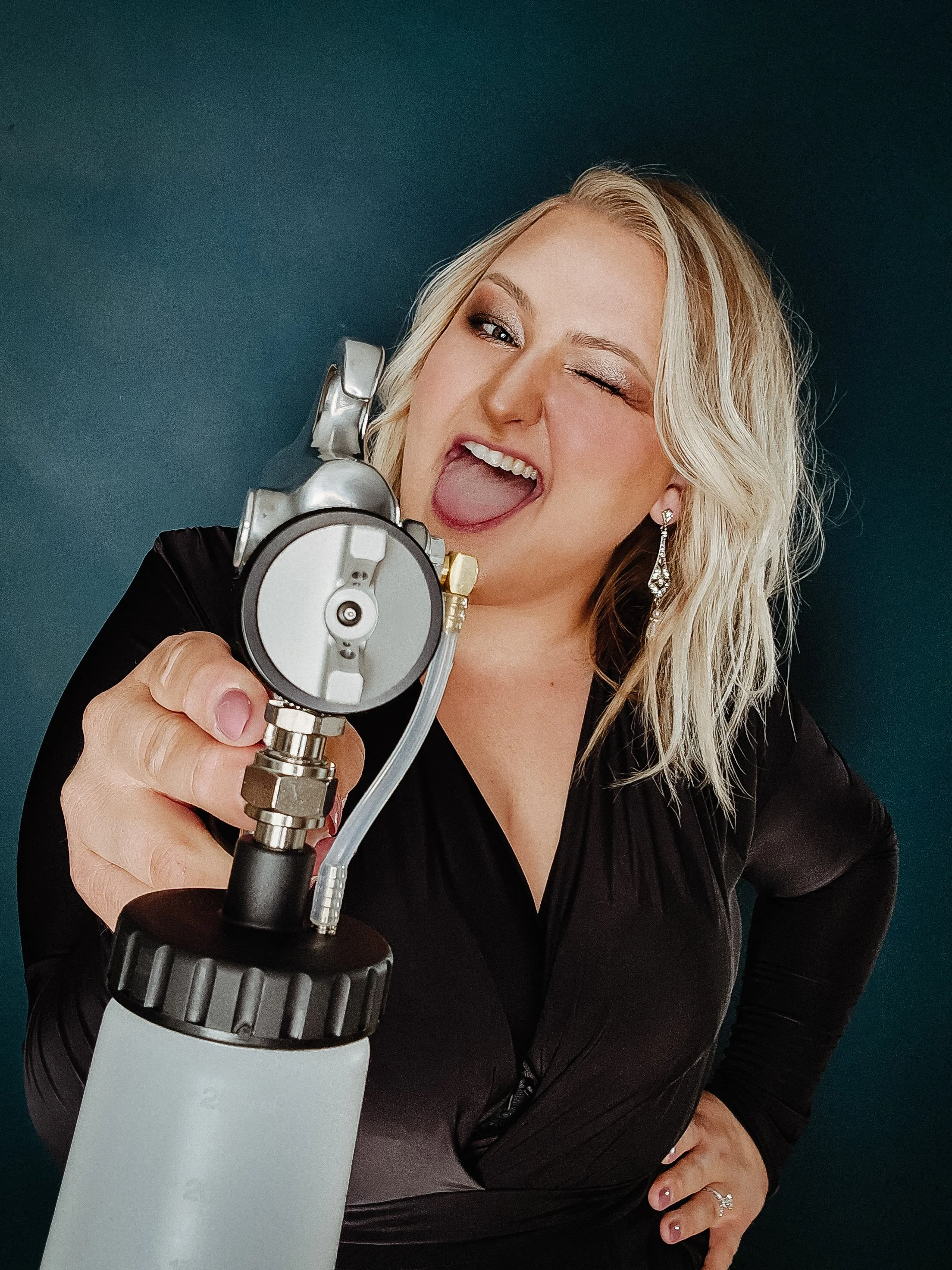 A woman with blonde hair wearing earrings and a black dress, holding a spray paint gun, this image is of the owner of Bronze Couture in Kalamazoo, Michigan that provides spray tanning and teeth whitening services.
