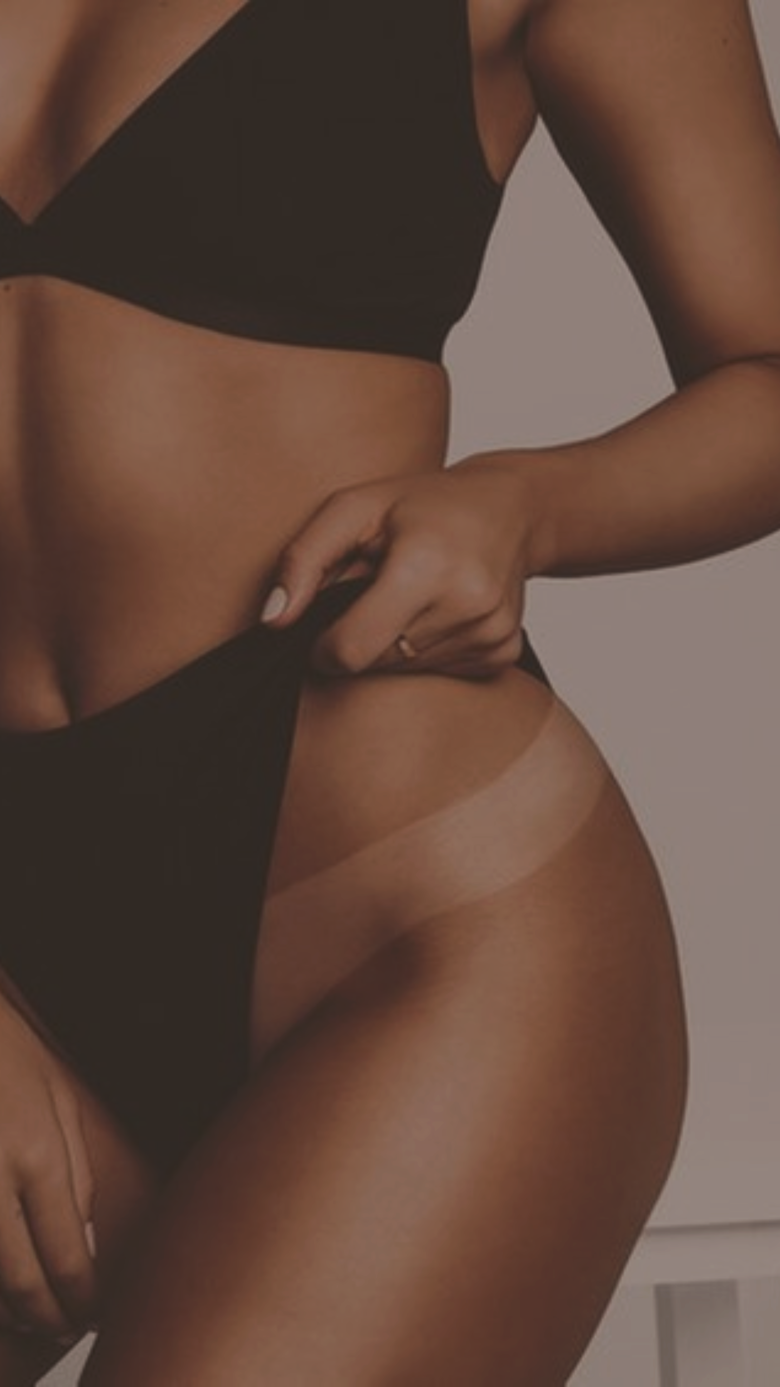 A woman is standing and wearing black underwear, pulling up the waistband of her underwear with her right hand, revealing her flawless spray tan from Bronze Couture in Kalamazoo, michigan spray tanning and teeth whitening.