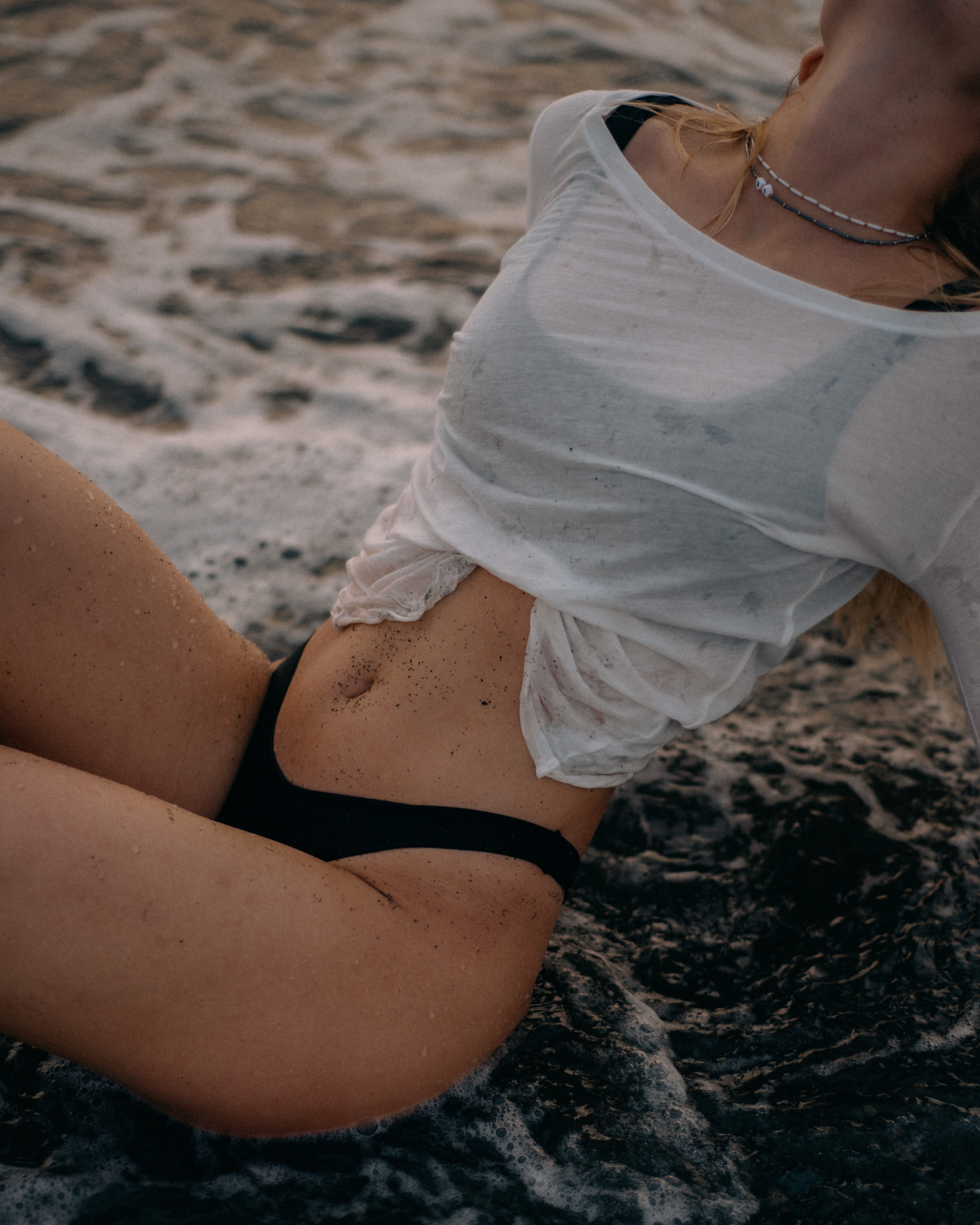 A woman in a white t-shirt and black underwear sitting on a sandy beach with water around her, with sand on her skin and a flawless spray tan at Bronze Couture in Kalamazoo, Michigan spray tanning and teeth whitening.