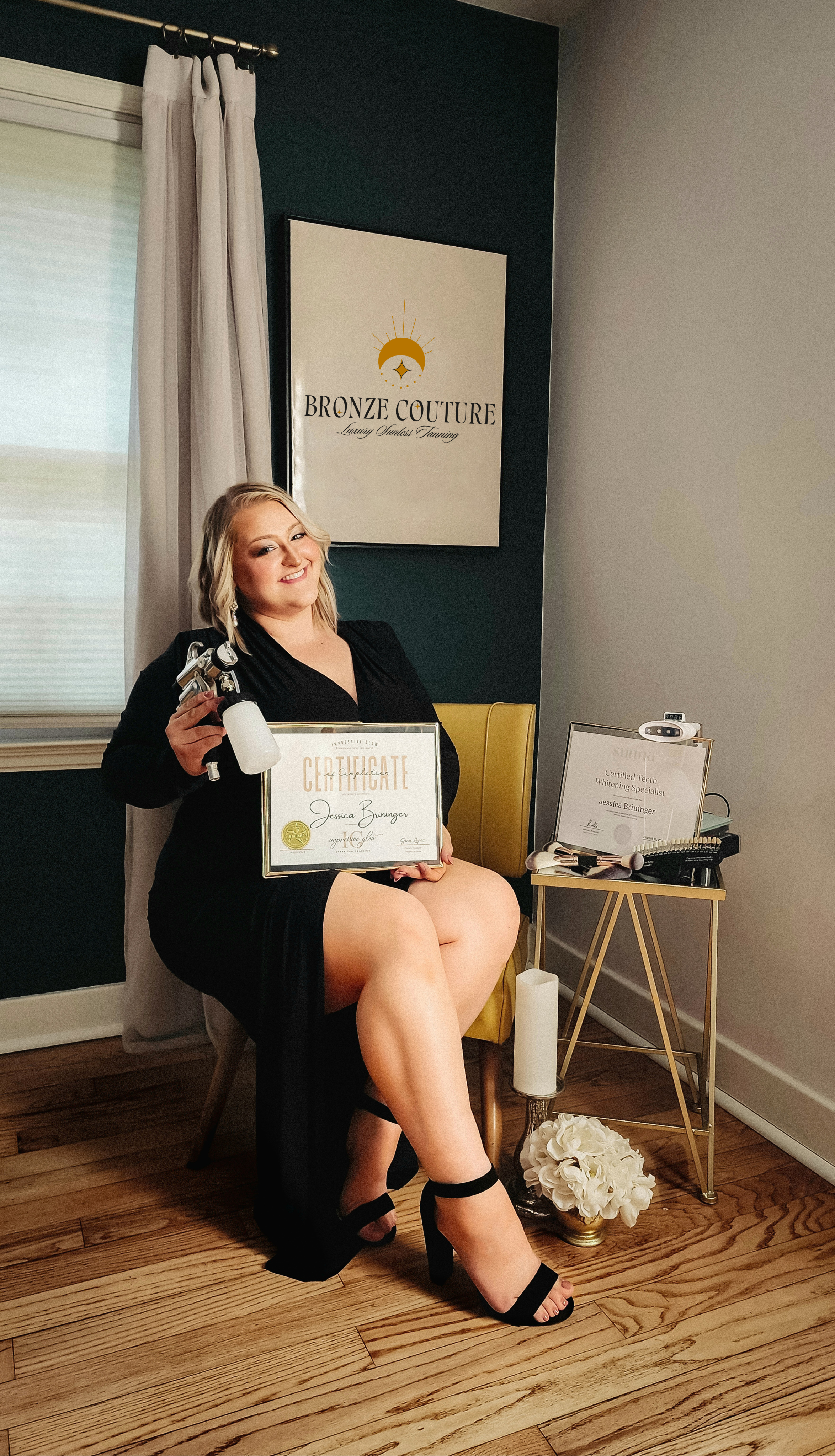 Woman in black dress sitting on yellow chair, holding a certificate, with a certificate on the table beside her, in a room with a sign that reads 'Bronze Couture' on the wall. This is the owner of Bronze Couture in Kalamazoo, Michigan spray tanning.