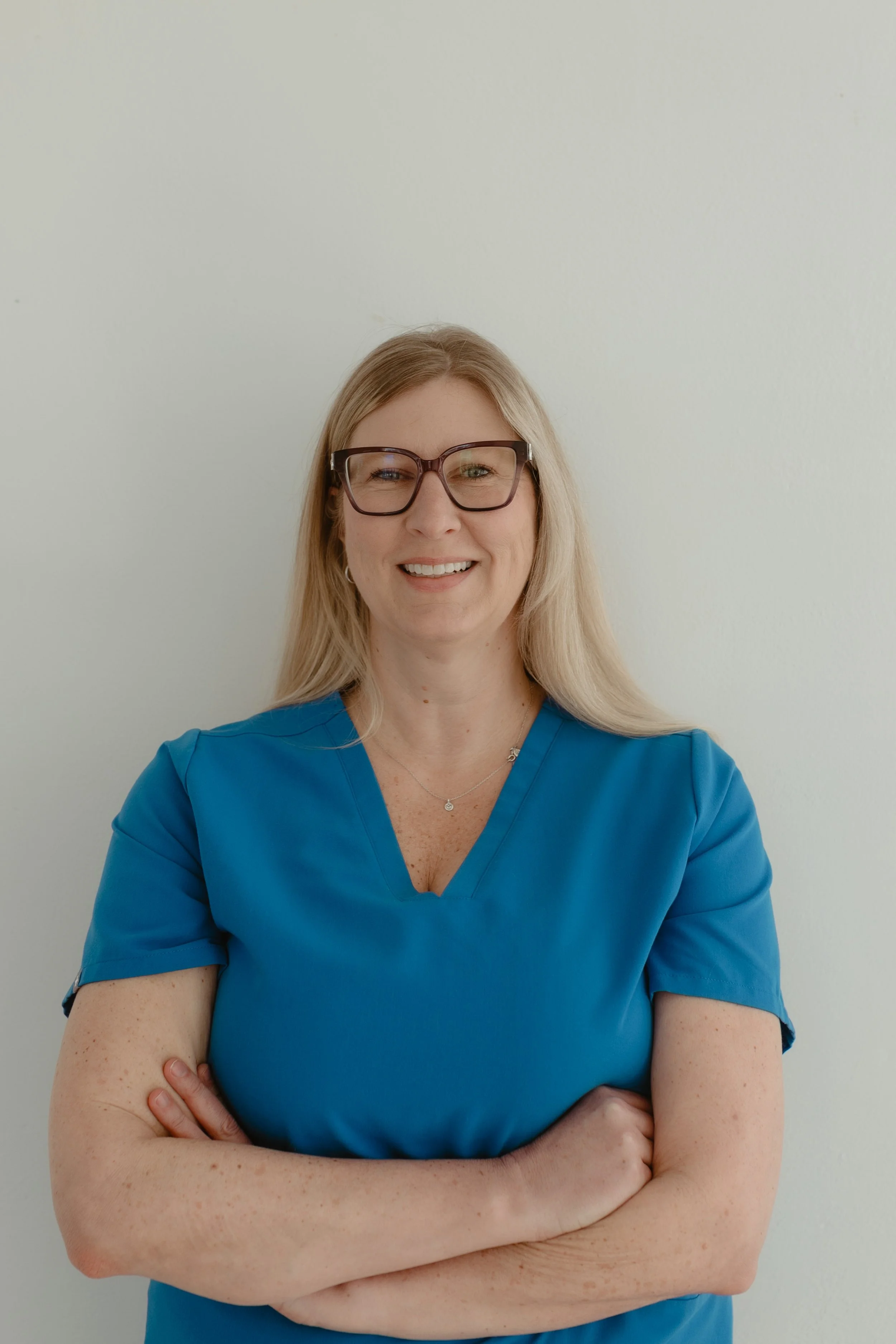 A woman with long blonde hair wearing glasses and blue scrubs, smiling and standing against a plain white wall.