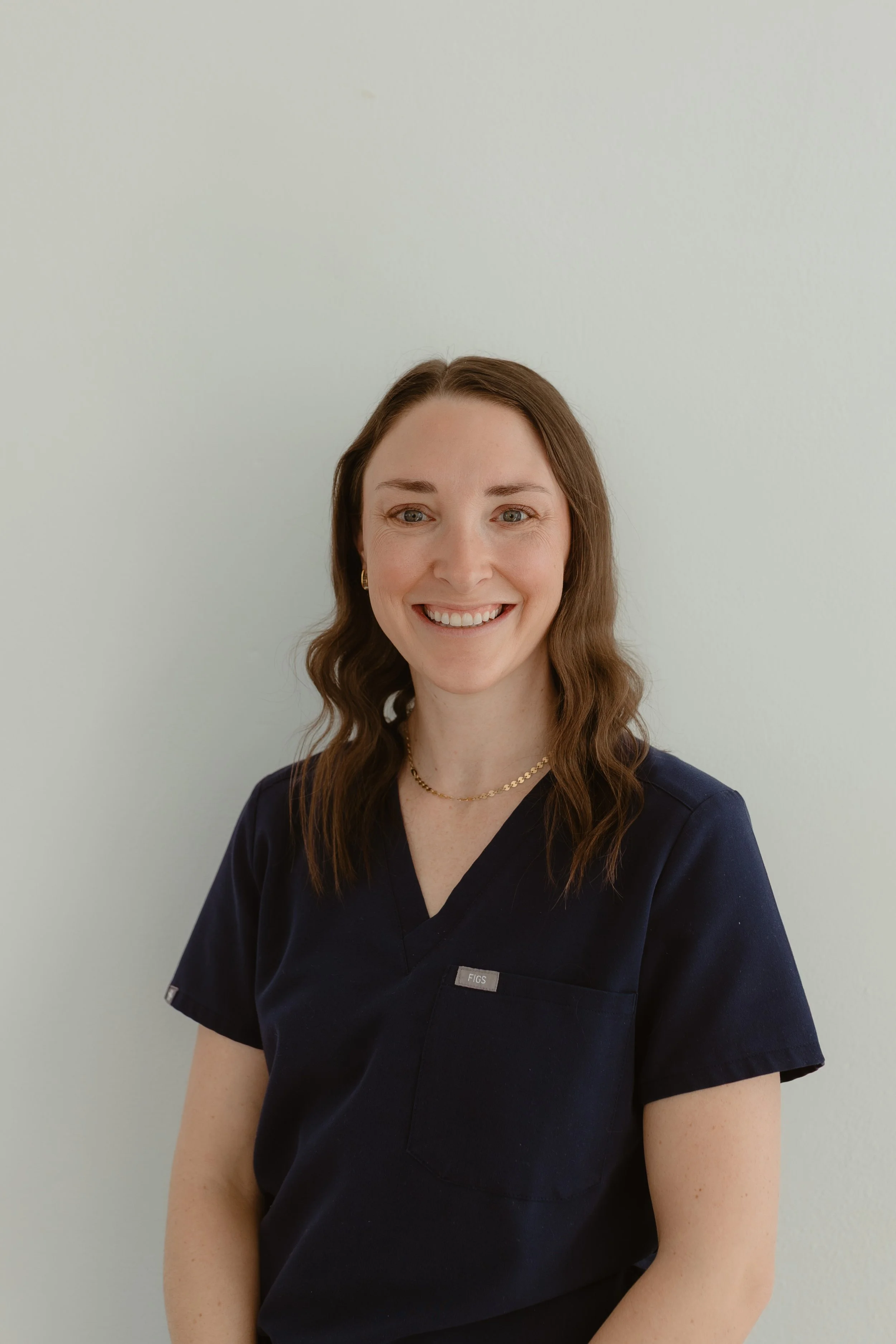 A woman with long brown hair, blue eyes, and a bright smile, wearing navy medical scrubs, gold earrings, and a gold necklace, standing against a white background.