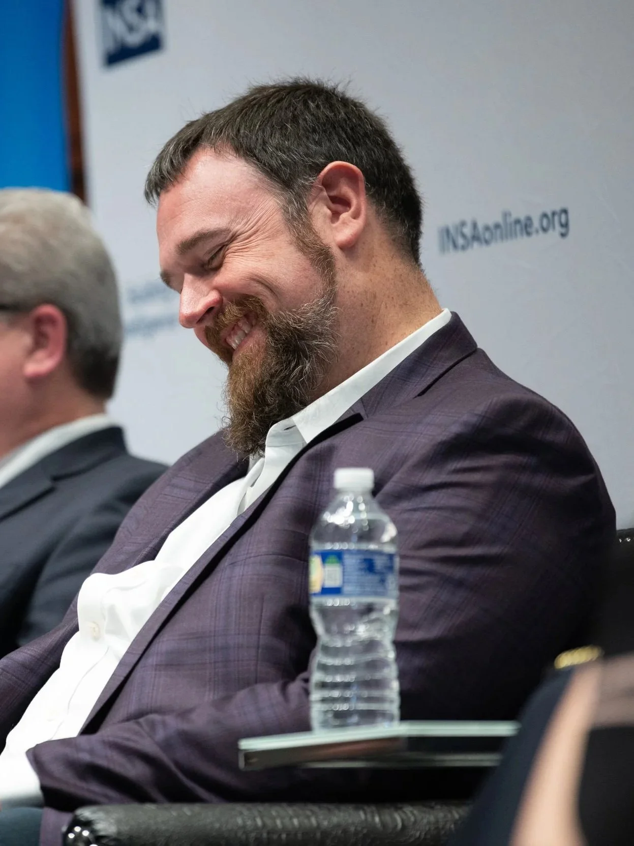 Brian Drake sitting at a conference panel discussion, he is smiling and looking down. Another man is reading a booklet. Both have water bottles in front of them, and a blue and white banner with the INSA logo is visible in the background.