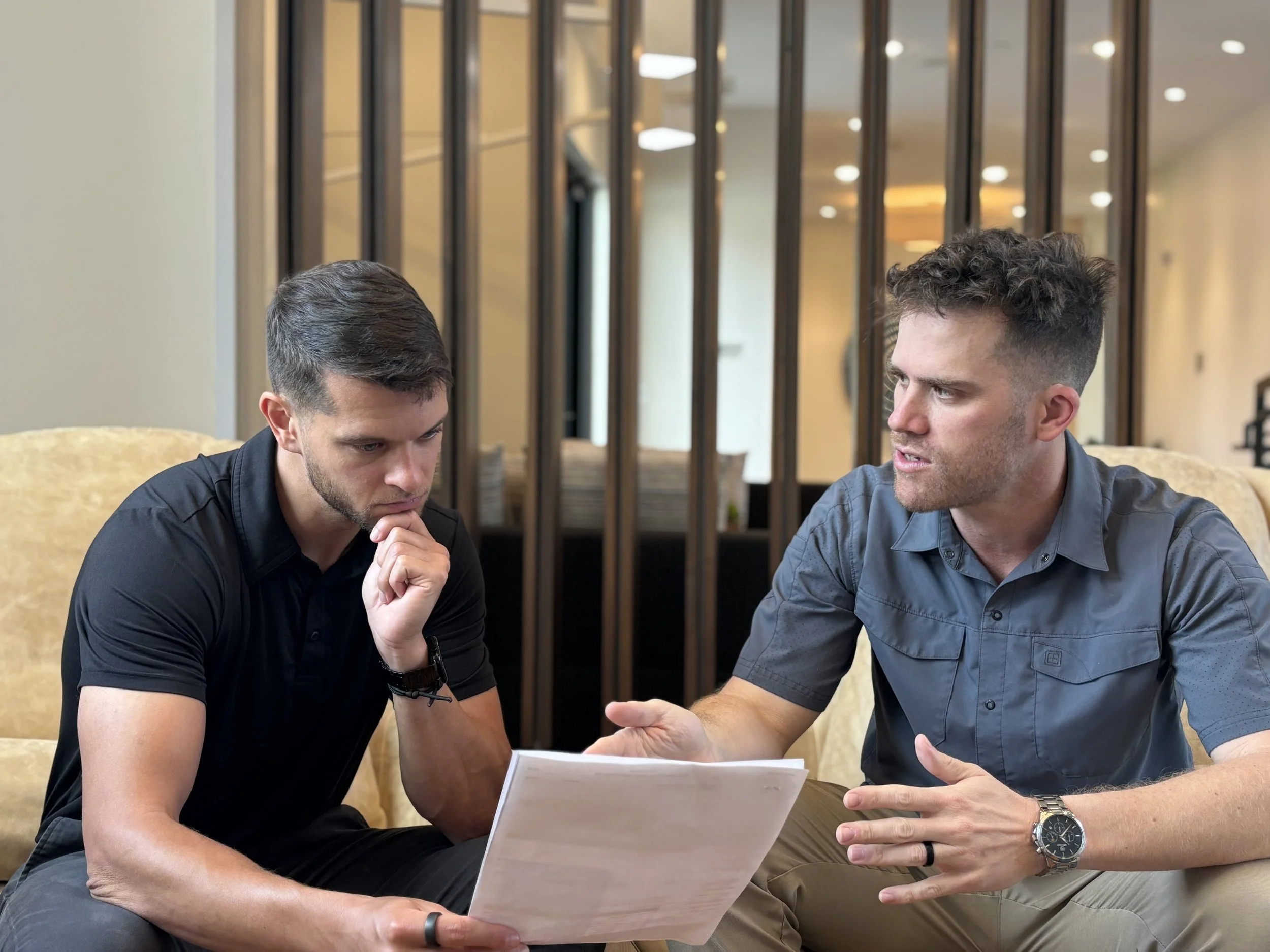 Two men engaged in a serious discussion, sitting at a table in a modern, well-lit interior with decorative wooden panels in the background. One man is holding a document.