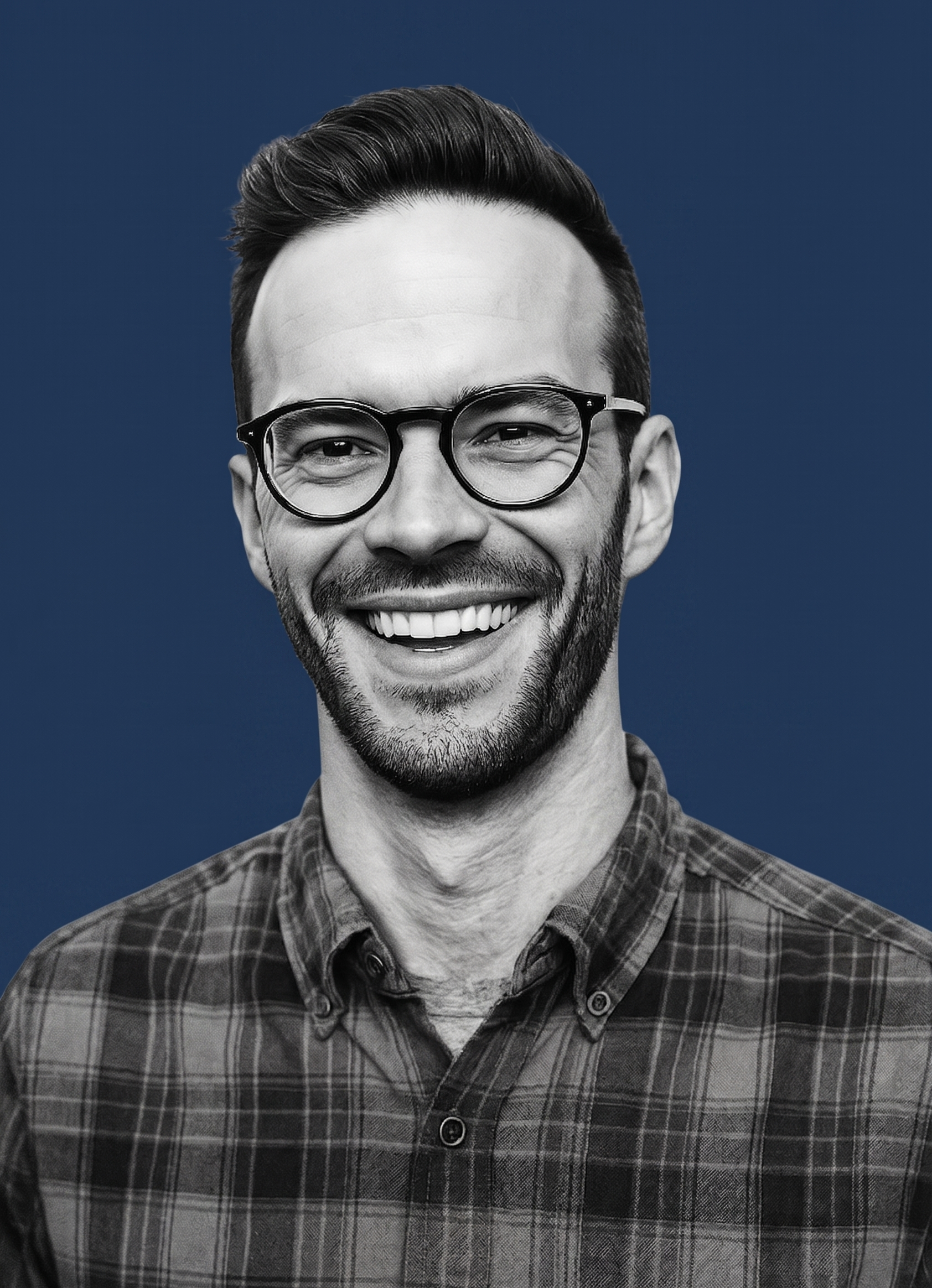 Black and white portrait of a smiling man with glasses, short dark hair, and a beard, wearing a plaid shirt, against a dark background.